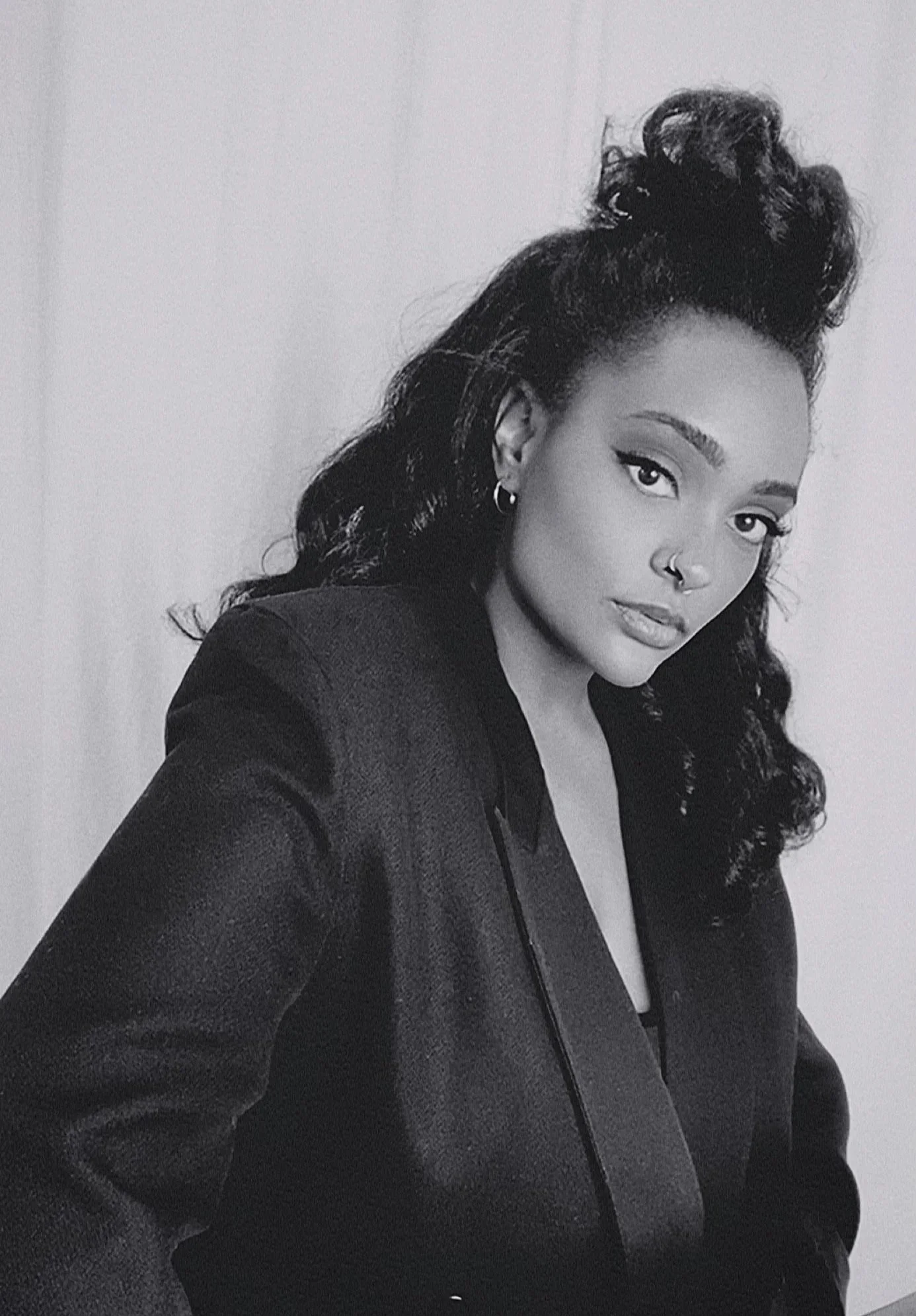 Black and white portrait of a woman with long curly hair styled in an updo, wearing a dark blazer, hoop earrings, a nose ring, and a small hoop in ear, looking directly at the camera with a confident expression.