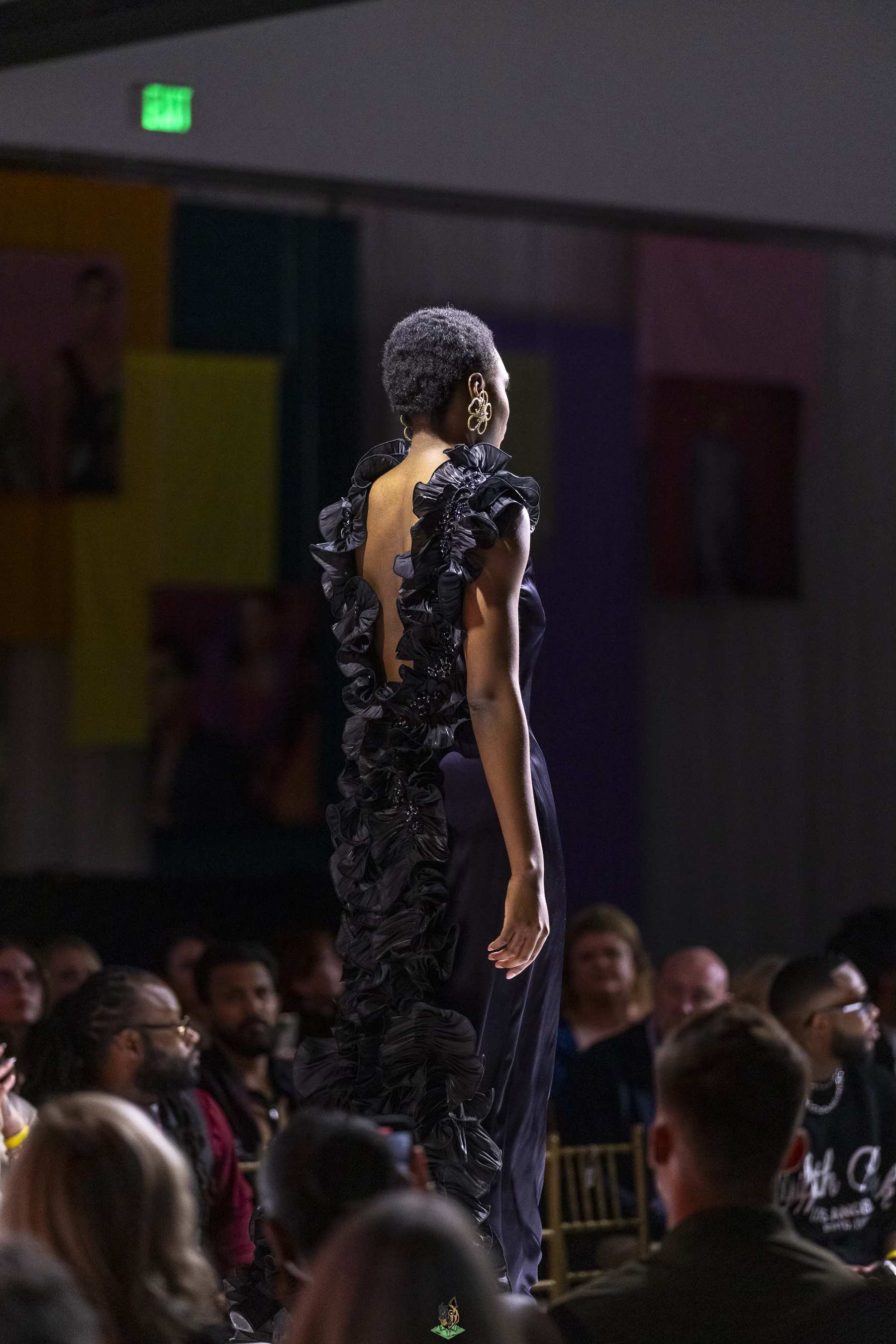 A runway model walking on stage during fashion show, wearing black ruffled gown with open back, large gold earrings, short natural hairstyle, audience seated on either side watching.