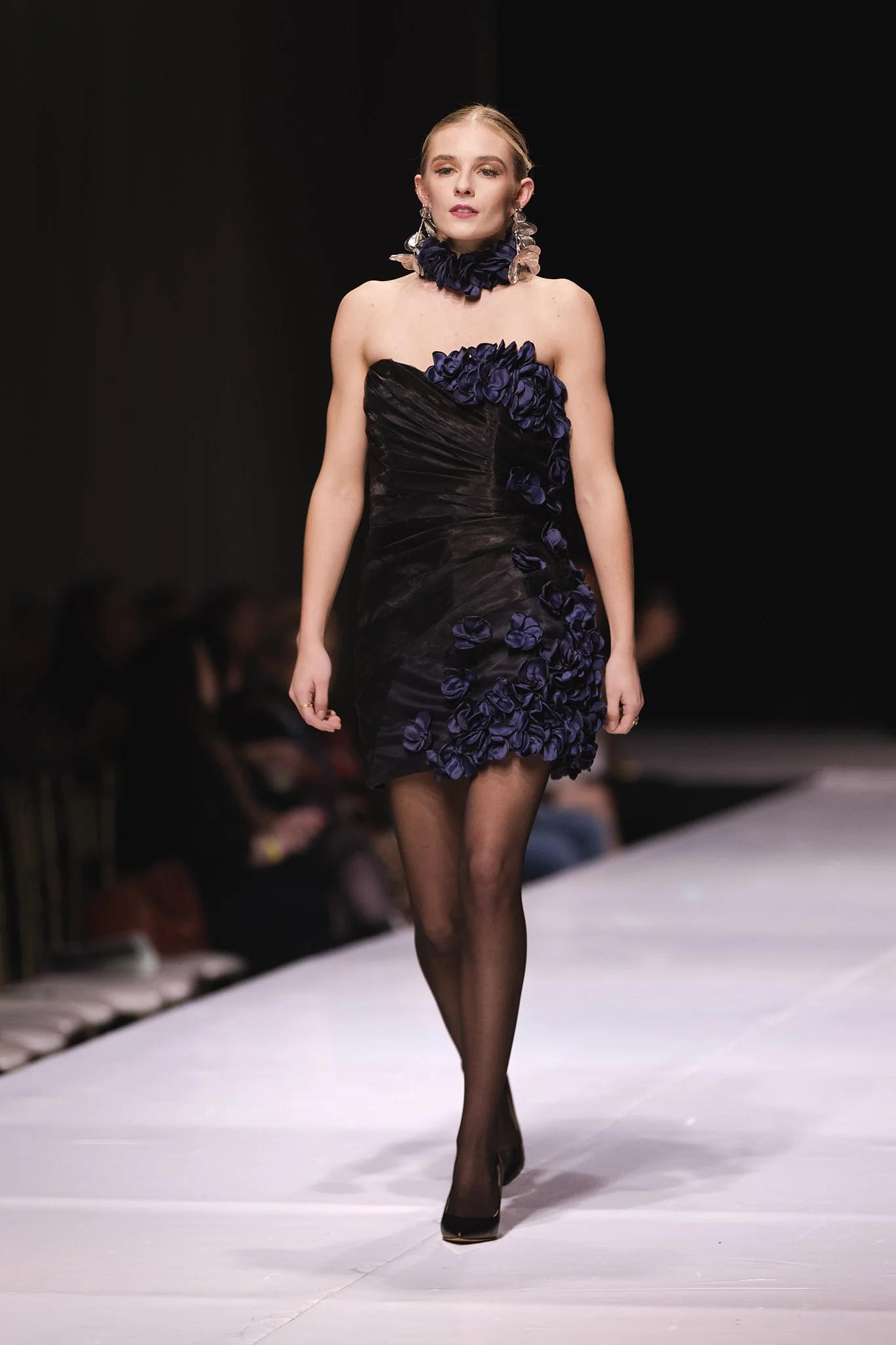 Female model walking on a runway, wearing a black dress with black flowers and a matching black floral choker, black stockings, and black heels.