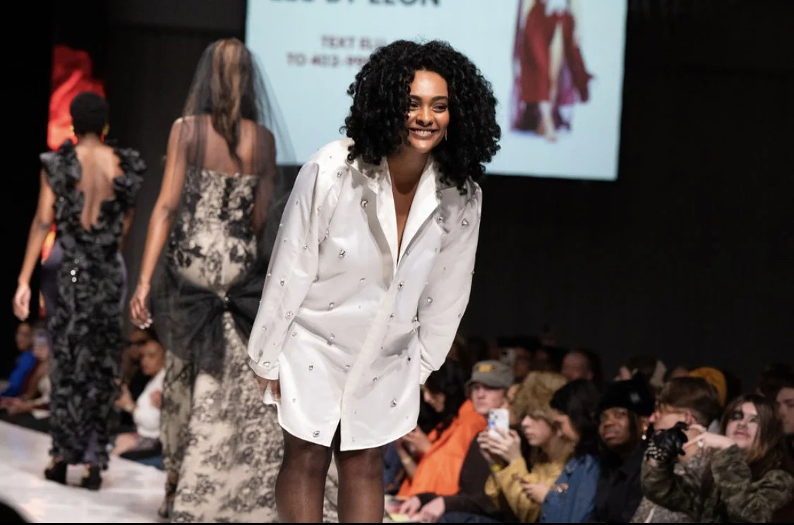 A woman with curly black hair smiling on a runway, wearing a white button-up dress with embellishments, with models in dresses walking behind her and an audience watching.