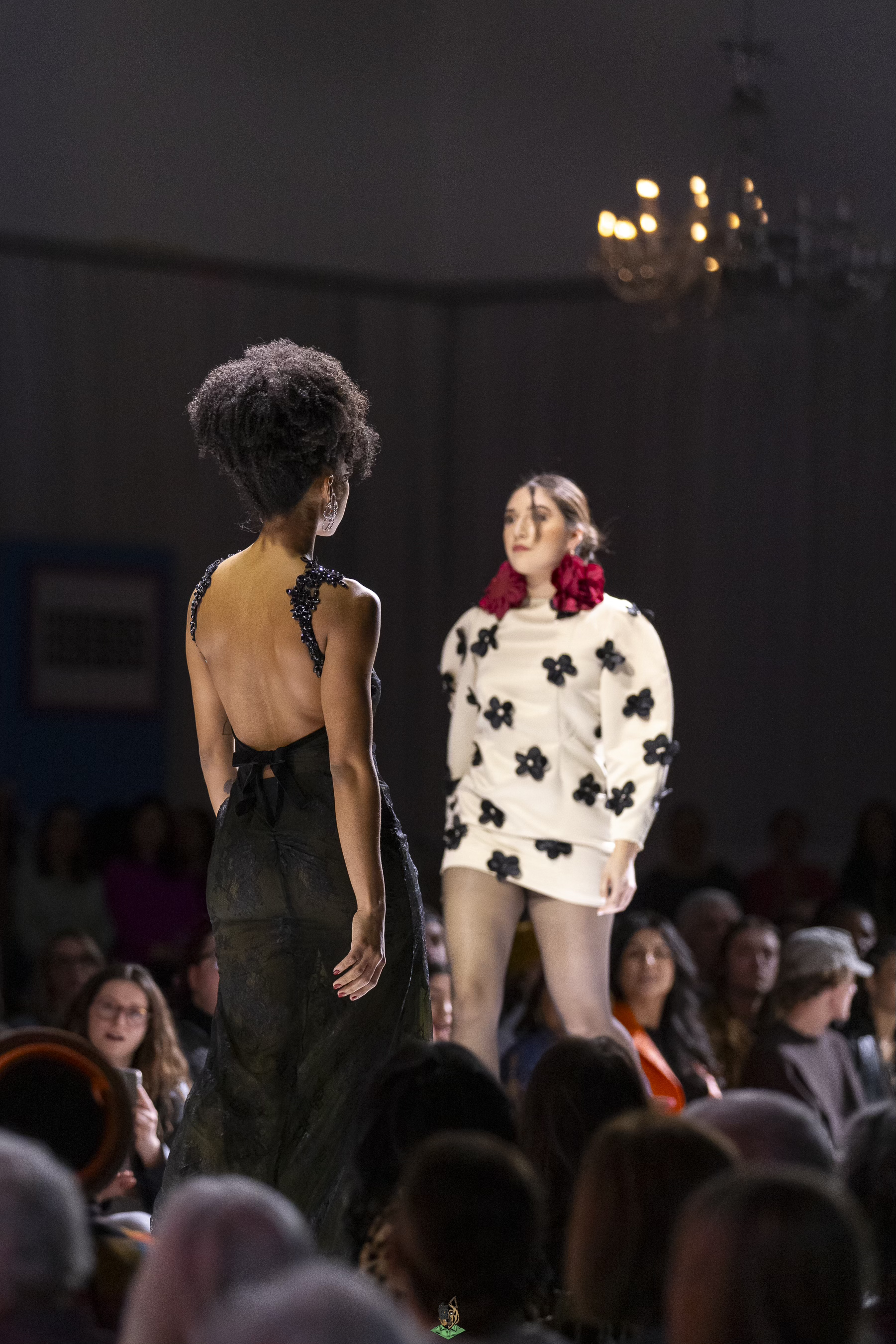 Two women walking on a runway during a fashion show, with the audience seated around them.