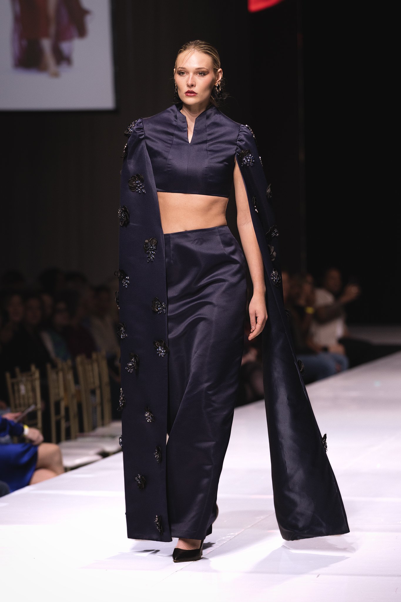 Model walking on runway wearing a navy blue two-piece outfit with a cape. The top is a cropped blouse with a high collar, paired with column skirt. The cape has decorative embellishments.