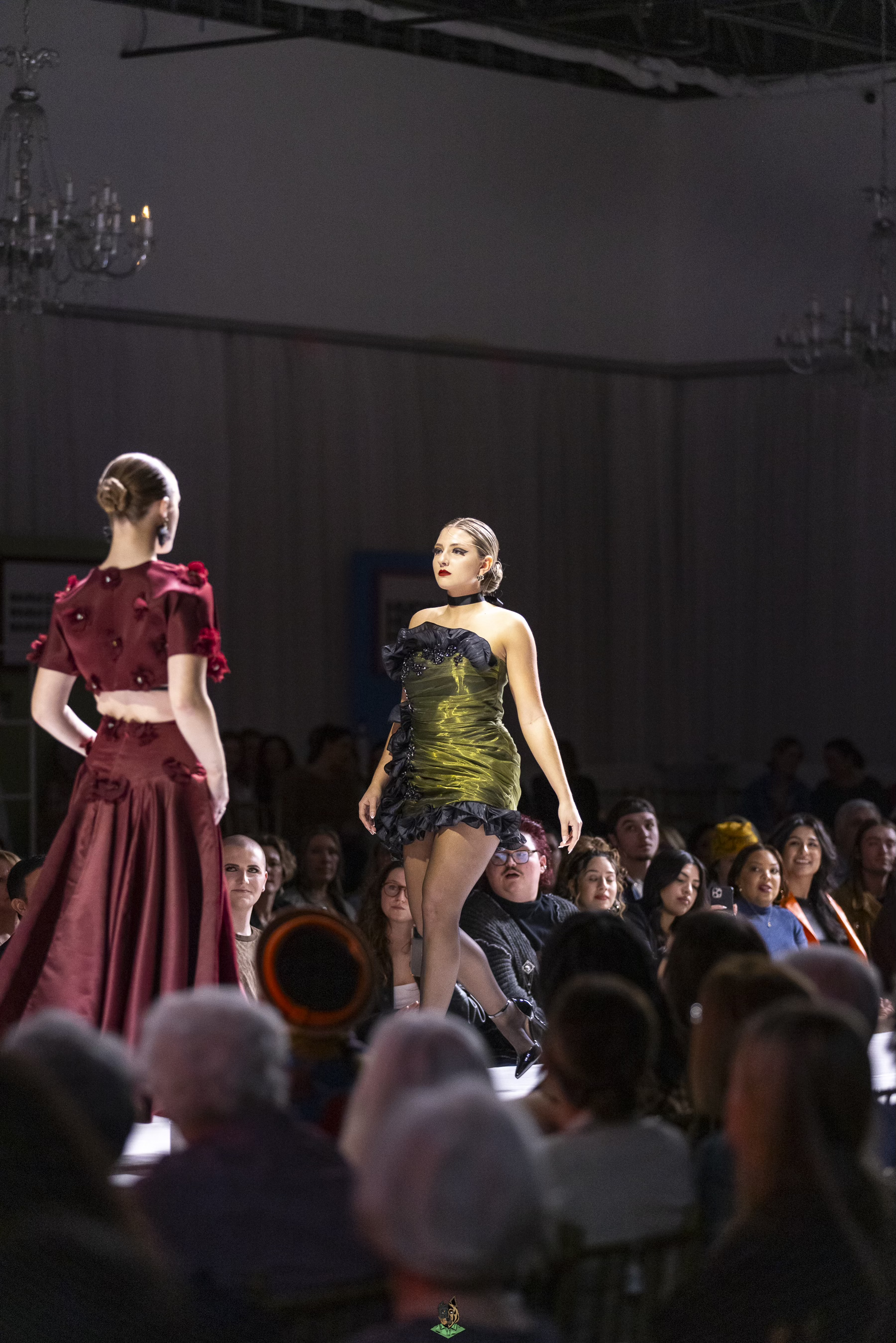 Two women walking on a runway during a fashion show, with an audience sitting on both sides of the runway, and chandeliers hanging from the ceiling.