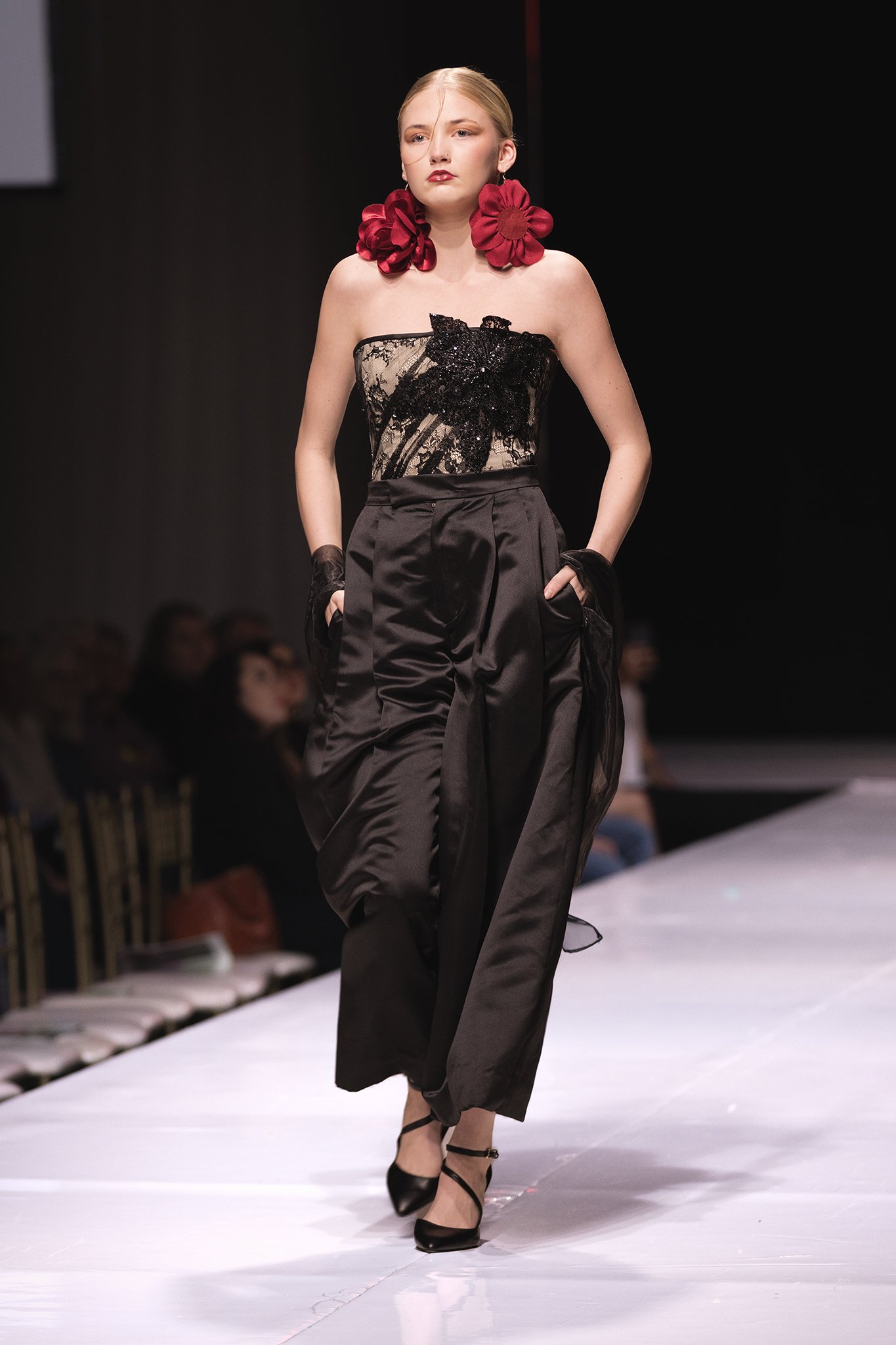 Model walking on runway wearing black satin wide-leg pants, a strapless lace top with black embroidery, red floral earrings, and black heels.