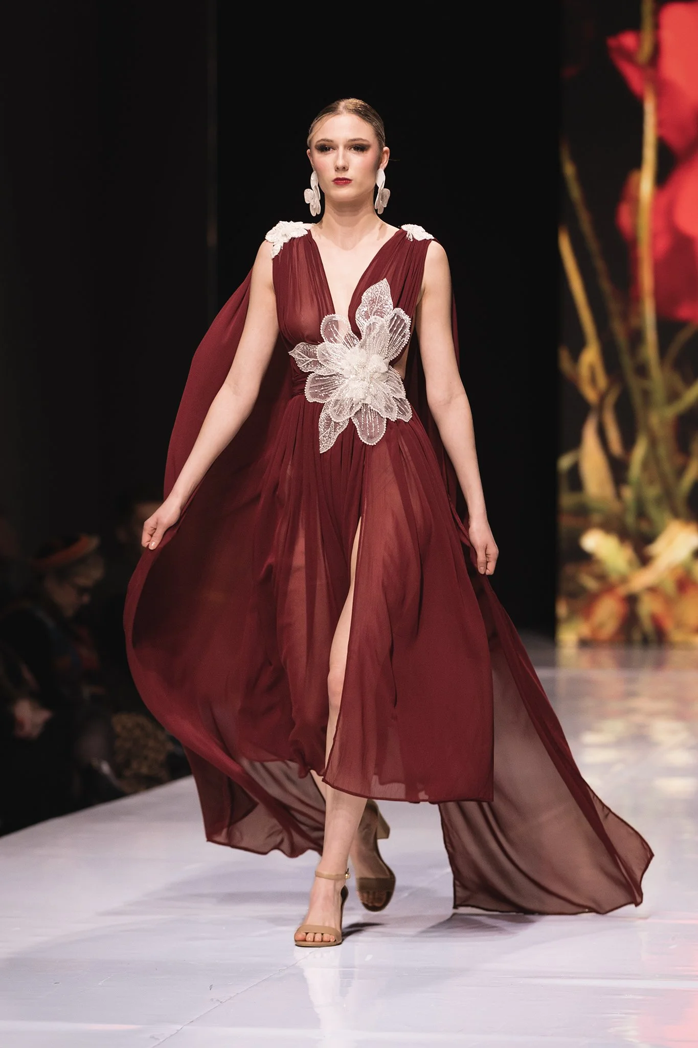 A female model walking on a runway in a burgundy sheer gown with silver floral appliqué and high slit, paired with beige high heels and large white floral earrings.