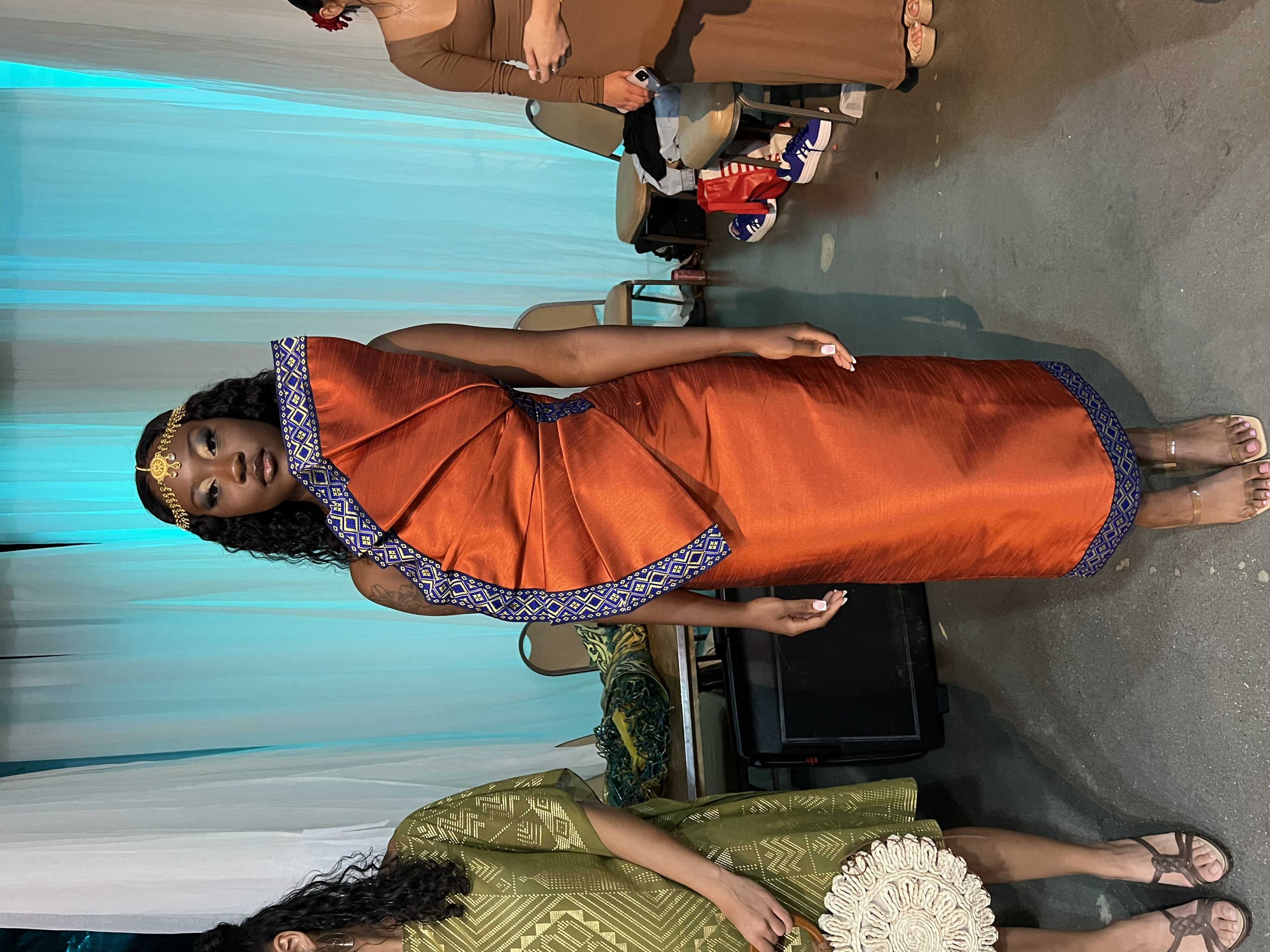 A woman dressed in a traditional orange dress with blue and gold accents, standing indoors near a table and chairs, with a turquoise curtain behind her.