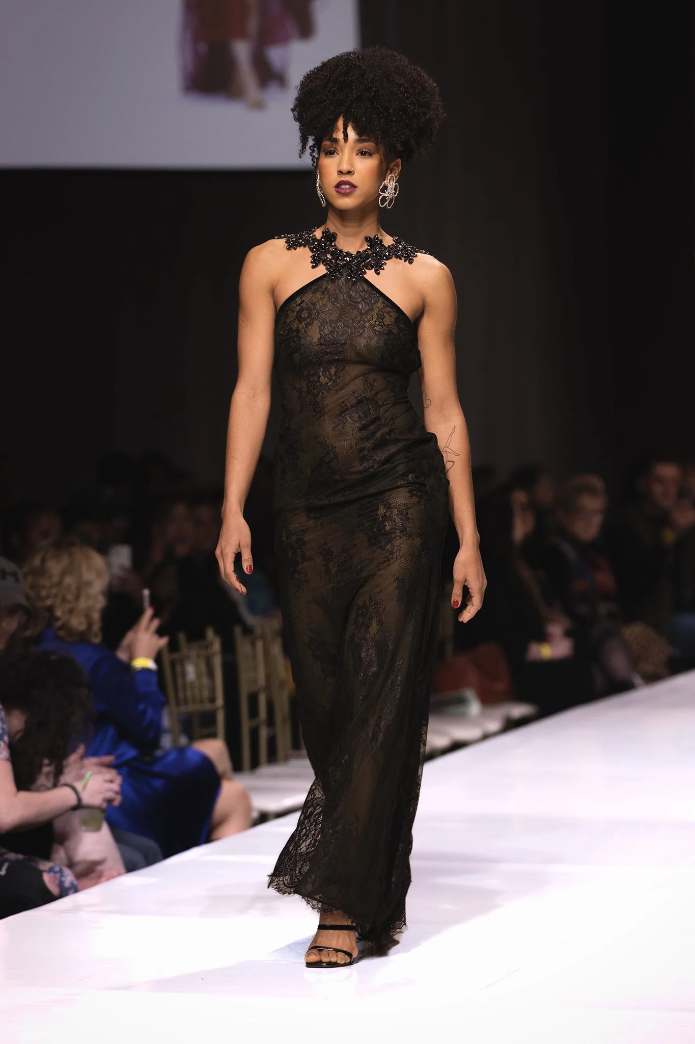 A woman walks on a runway during a fashion show wearing a black lace dress with intricate details and large earrings.