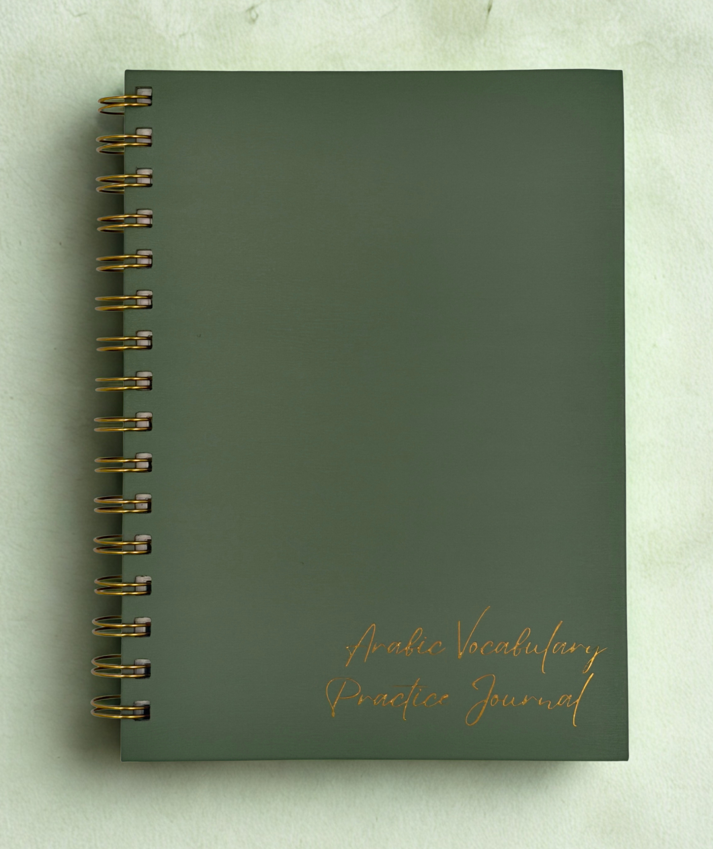 A green spiral-bound notebook with gold-colored rings, lying on a light green surface. The cover has gold cursive text that reads "Arabic Vocabulary Practice Journal".