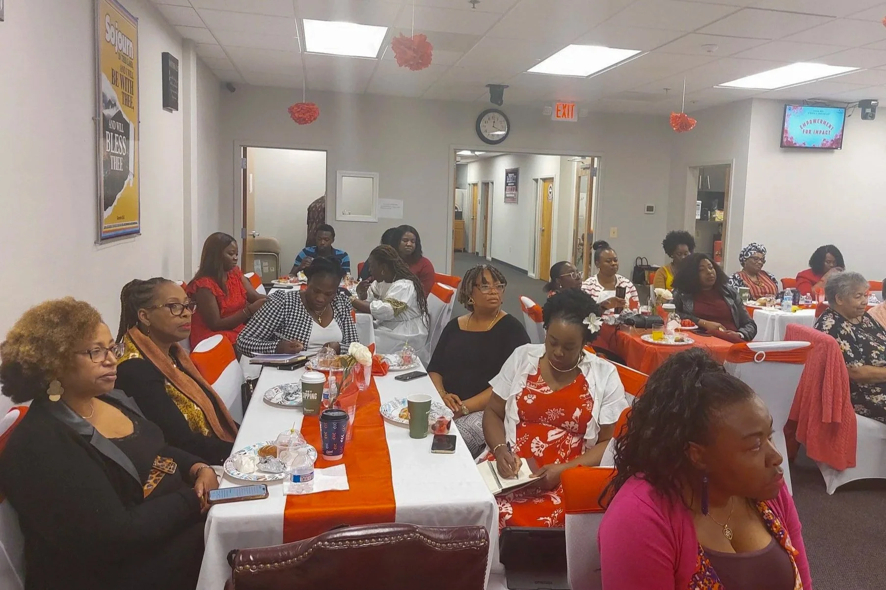 Women attending a meeting or event in a conference room with tables decorated with orange and white tablecloths. The women are seated, some taking notes or using their phones, and appear engaged. The room has posters on the walls and a clock showing 12:19.