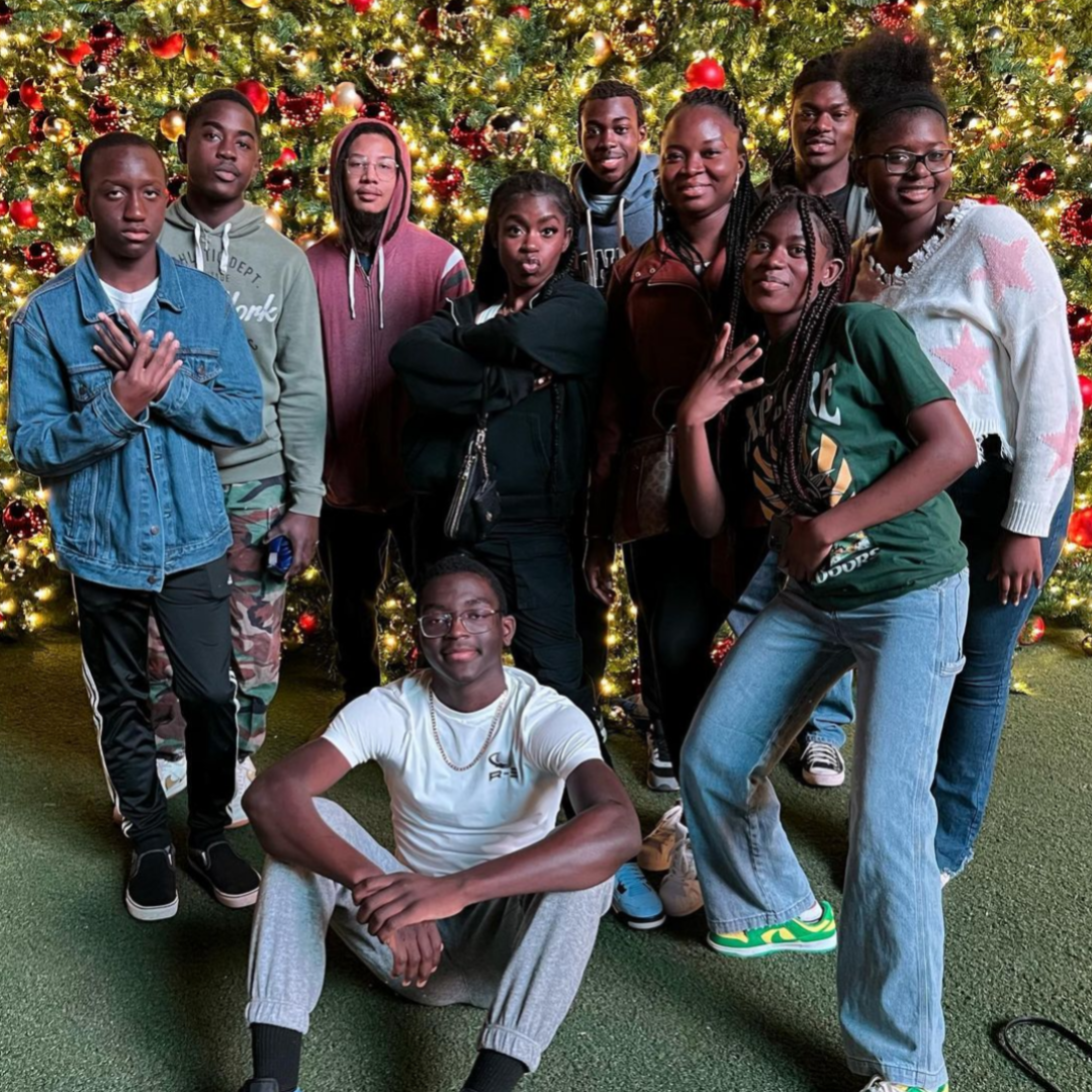 A group of nine diverse young people posing in front of a large decorated Christmas tree with red and gold ornaments and lights.