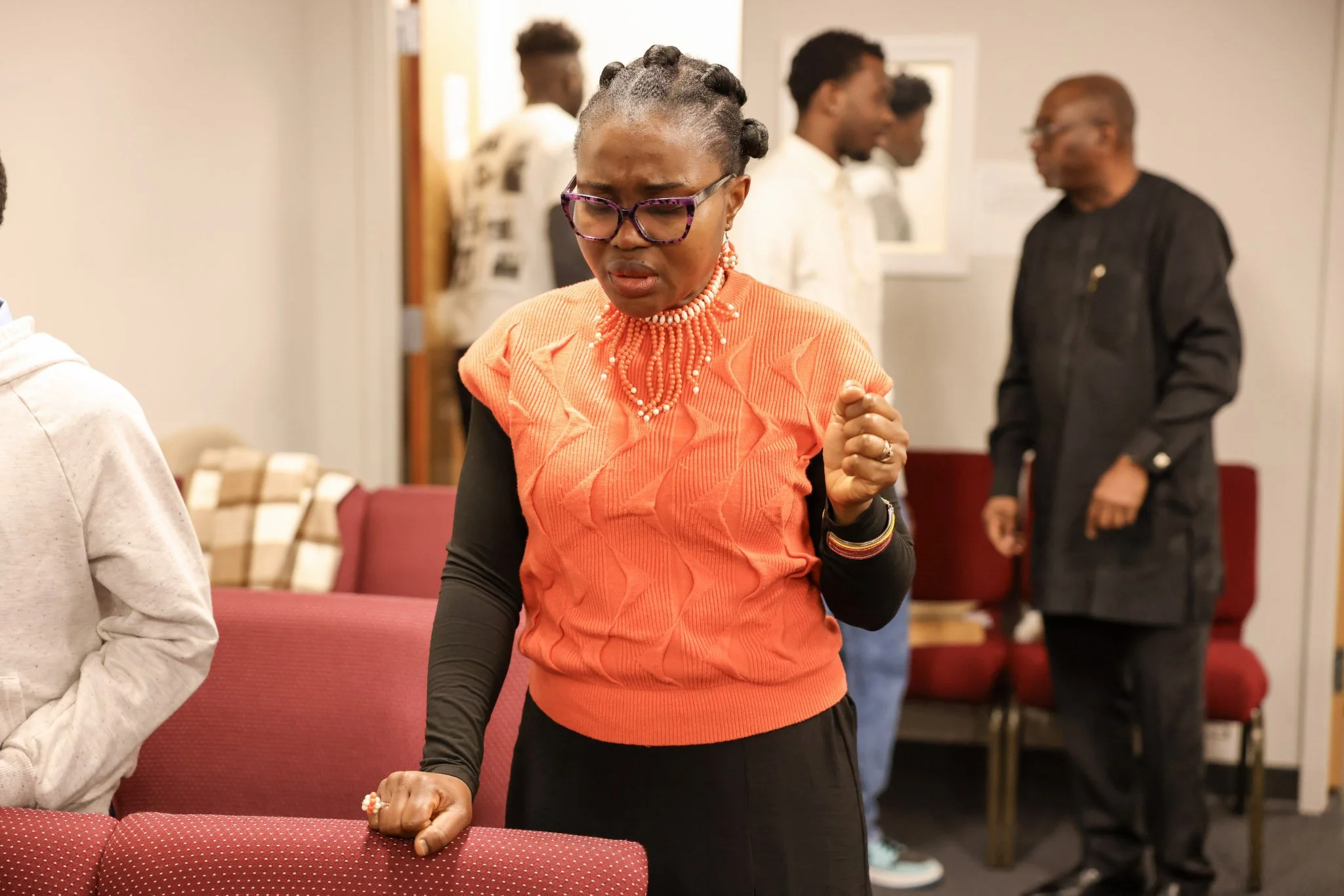 A woman with braided hair, glasses, Hebrew necklace, and an orange patterned top, appears to be praying or reflecting with her eyes closed and hand clenched in front of her, in a room with several people.