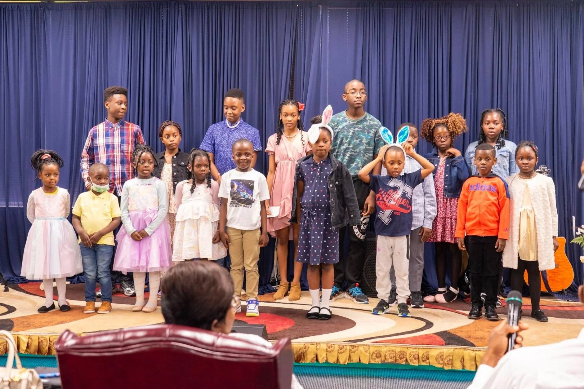 Group of children standing on stage with blue curtains in the background, some wearing bunny ears, during a performance or presentation.
