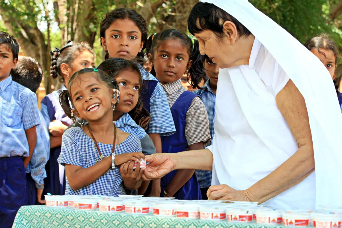 A young girl in school uniform smiling as she receives a gift from an elderly woman in white clothing, surrounded by other school children outdoors.