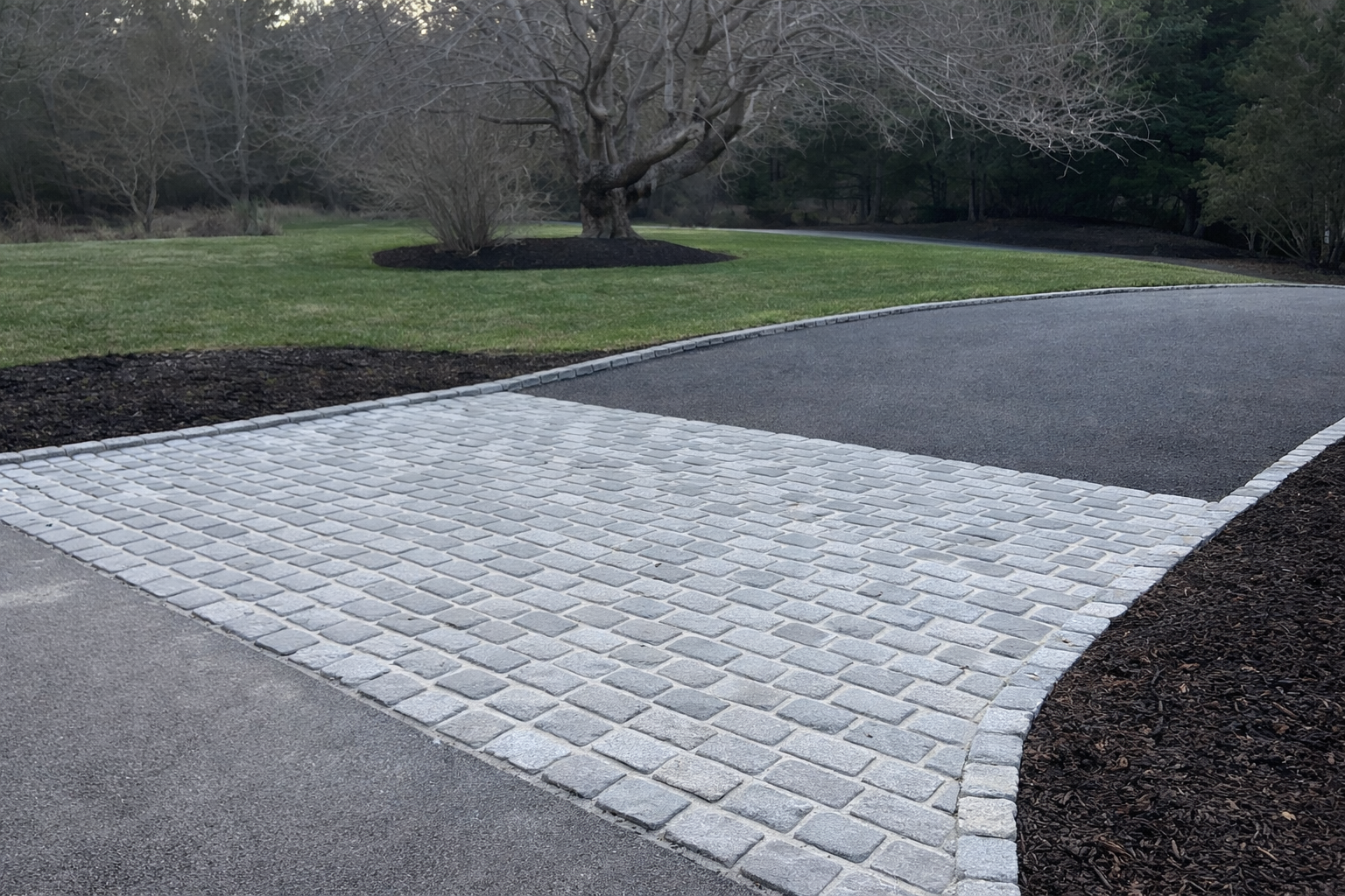 A paved driveway with gray cobblestones, bordered by dark soil mulch, leading to a curved asphalt road in a yard with a large leafless tree, green grass, and trees in the background.