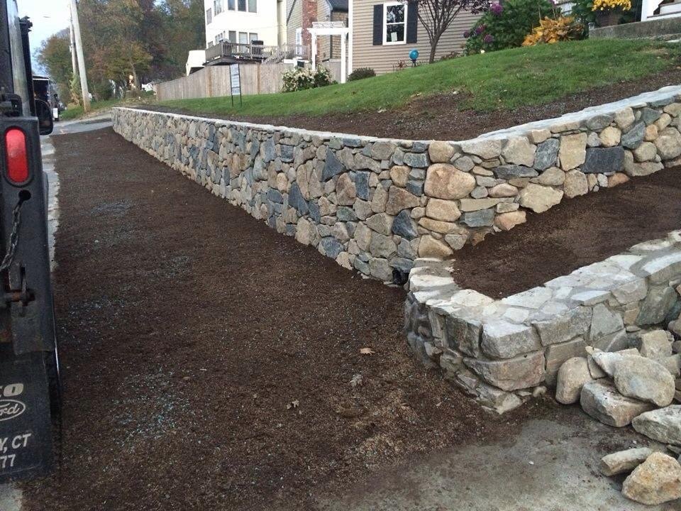 A stone retaining wall along a sloped yard, with a walkway on the side, under development.