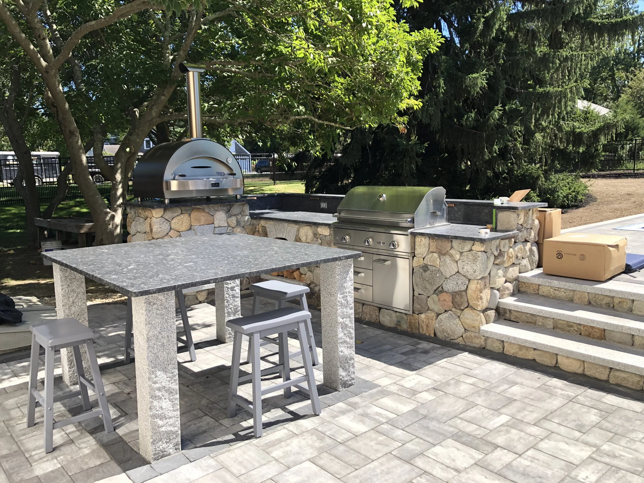 Outdoor kitchen area with stone countertops, a built-in grill, a pizza oven, and a table with stools, set on a paved patio with trees and greenery in the background.