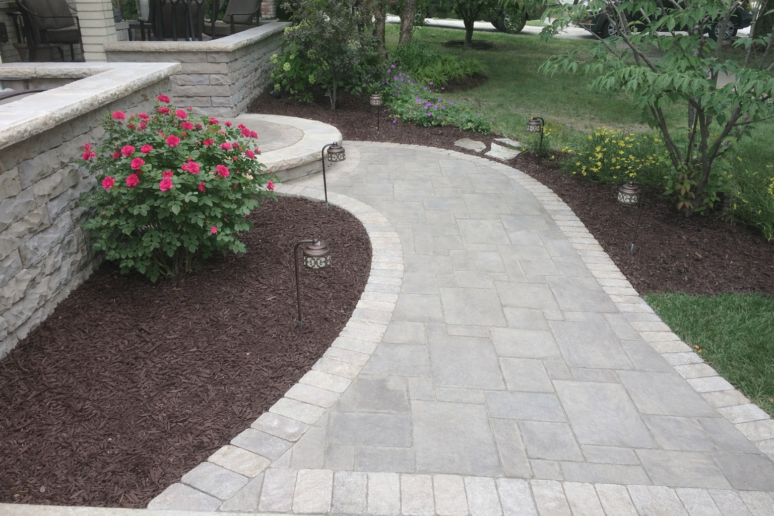 A curved stone walkway bordered by small stone edging, with a garden bed filled with mulch and blooming pink and purple flowers, surrounding a stone retaining wall with outdoor chairs and a patio in the background.