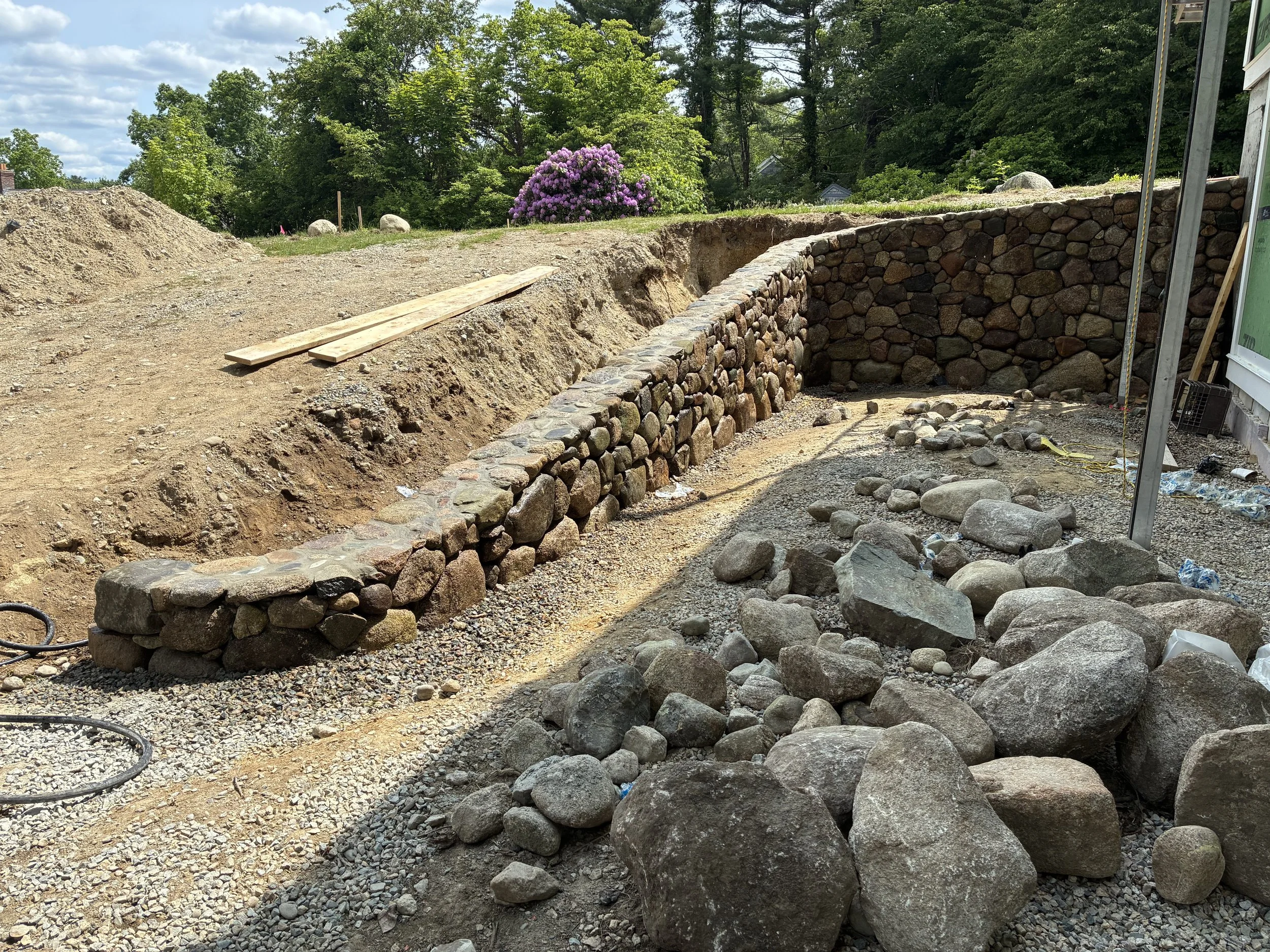 Construction site with a stone retaining wall and piles of rocks, dirt, and gravel, surrounded by trees and a flowering shrub in the background.