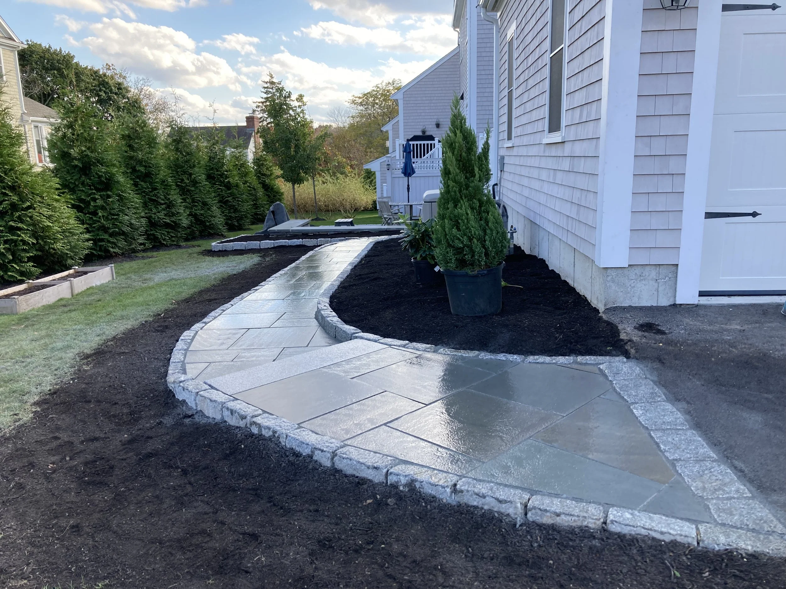 Newly paved winding concrete walkway in backyard with landscaped edges, green trees, and a house with a white garage door in the background.