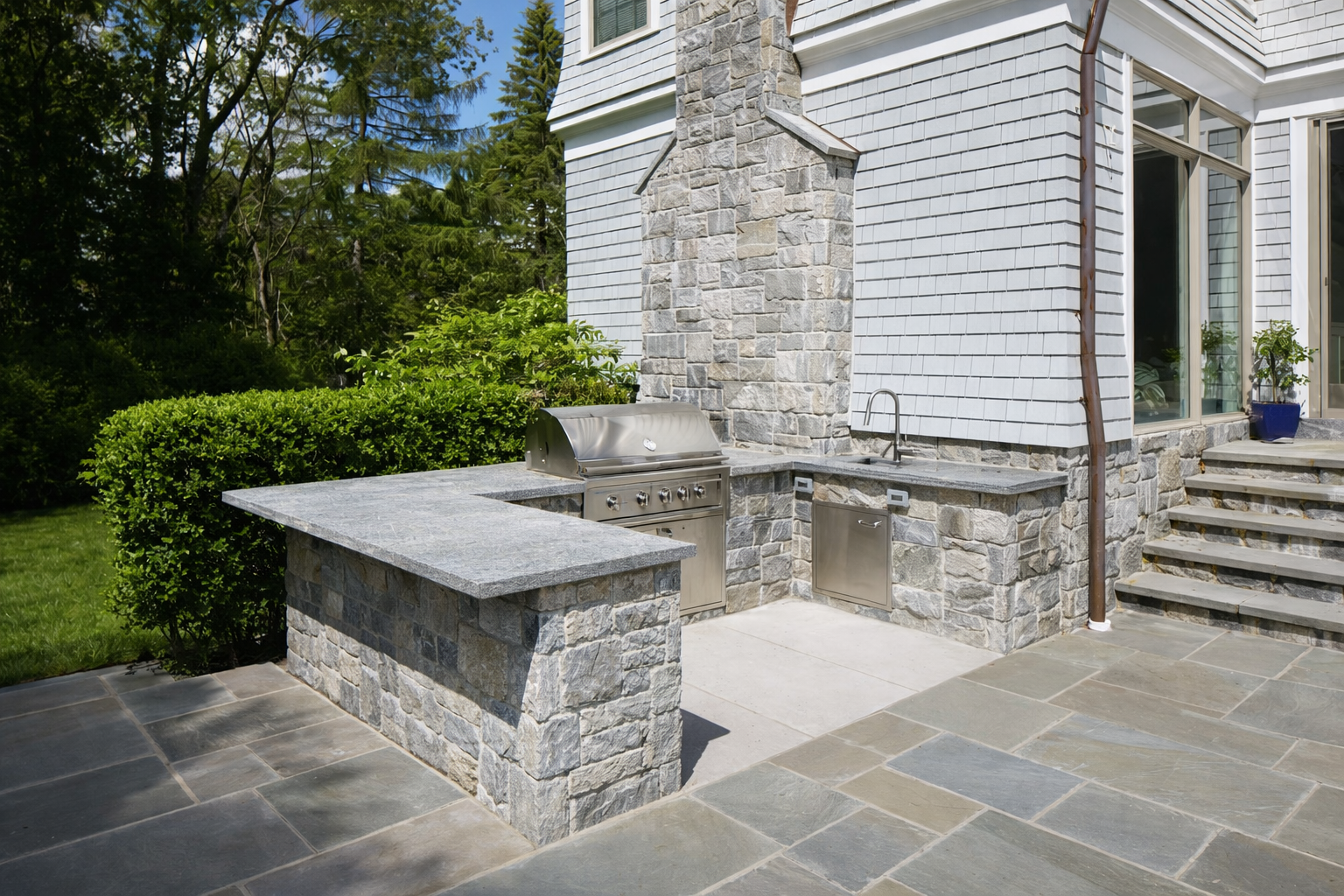 Outdoor backyard area with a built-in stone and granite BBQ grill, a sink, and countertops, next to a house with white shingle siding and stairs leading to a patio.