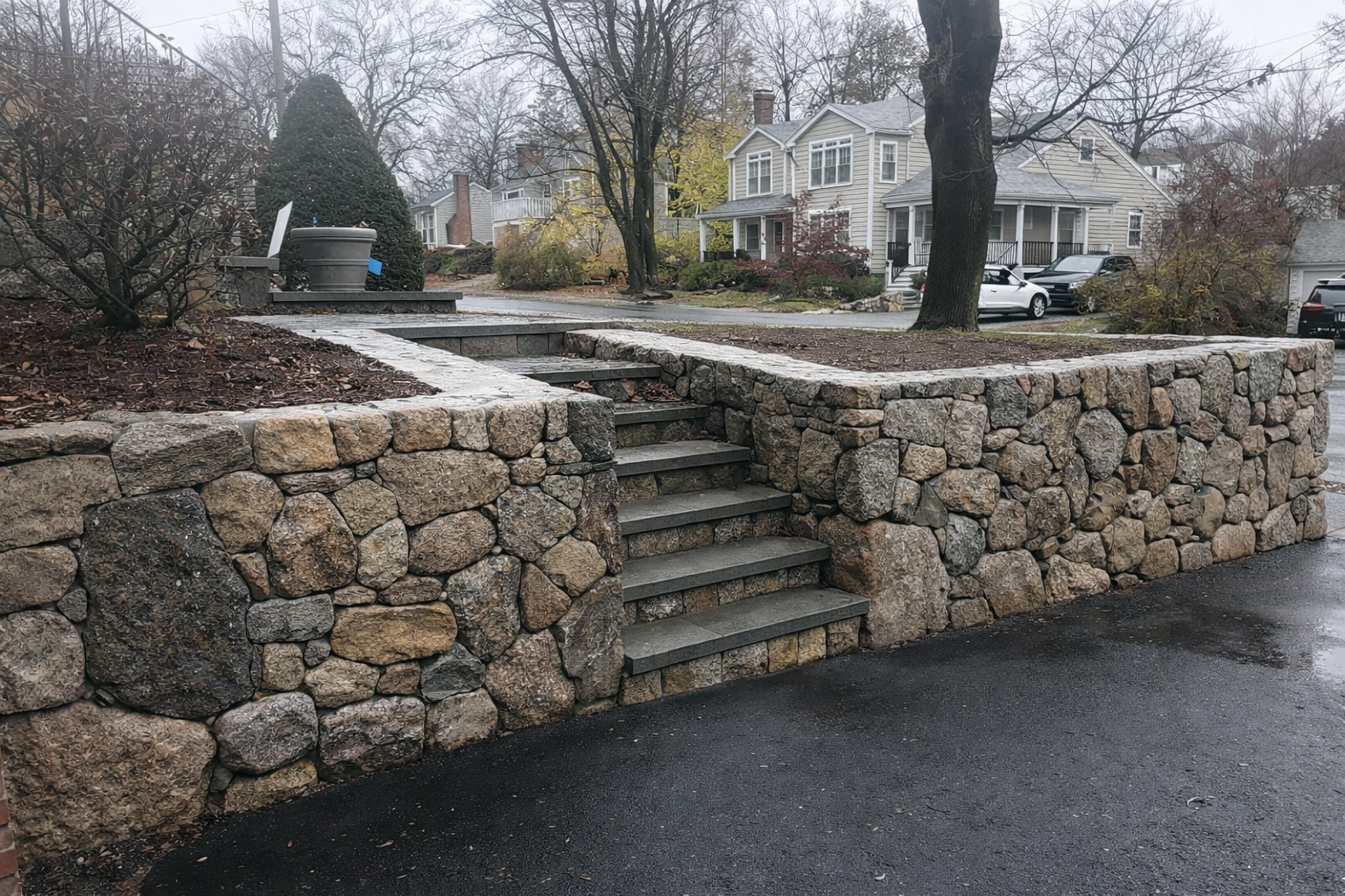 A stone retaining wall with steps leading up to a landscaped yard in a residential neighborhood, with houses, trees, and parked cars visible in the background on a cloudy day.