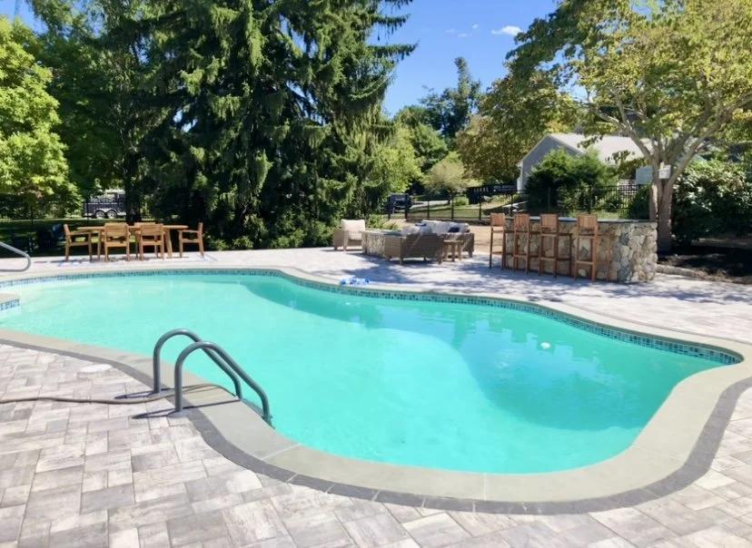 Empty backyard swimming pool with a metal handrail, surrounded by stone and tile paving. In the background, there are outdoor chairs, sofas, and bar stools near trees and a house, under a blue sky with some clouds.