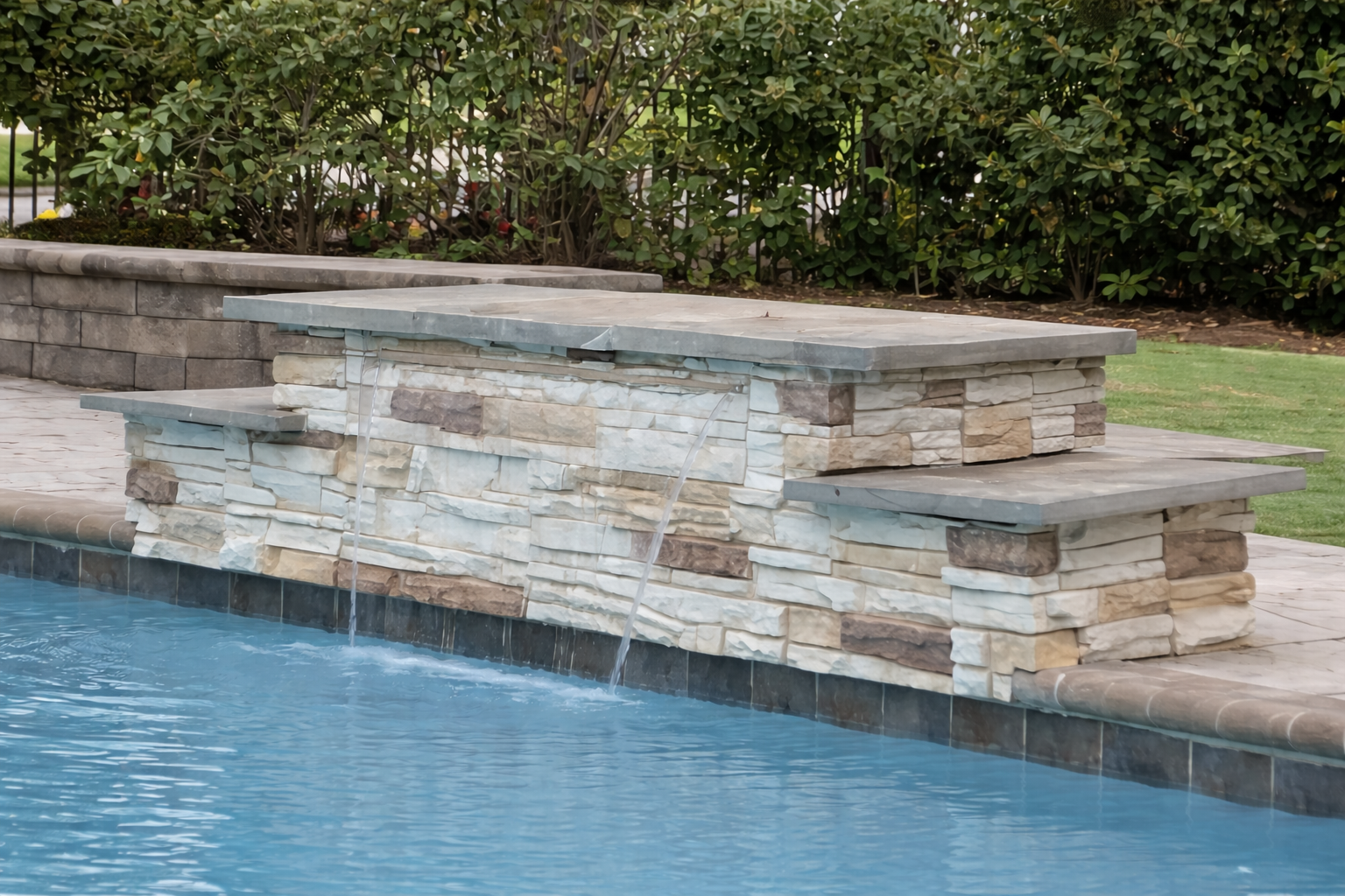Close-up of a stone pool fountain with water flowing from two spouts, surrounded by a stone patio and lush greenery.