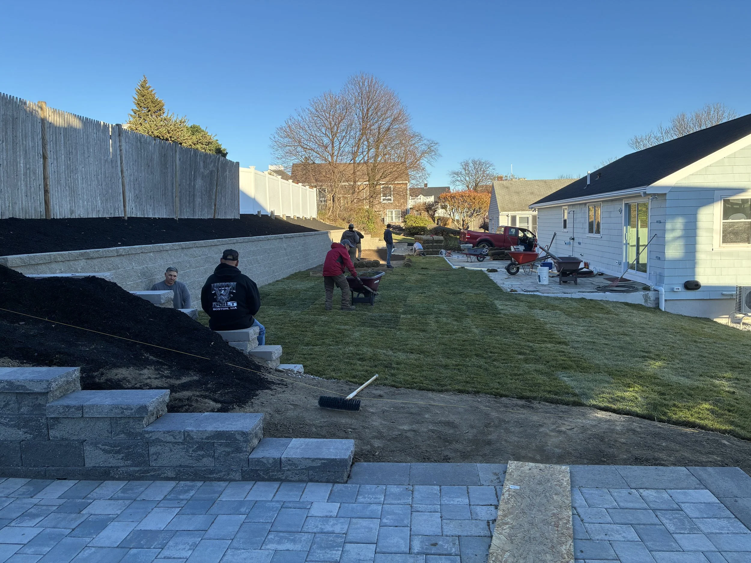 People working on a backyard landscaping project, installing retaining walls, laying paving stones, and gardening as part of outdoor renovation on a clear, sunny day.