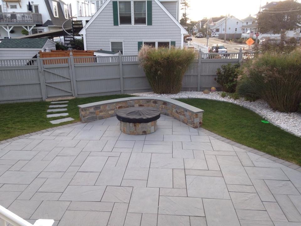 Backyard patio with large tiled surface, stone fire pit with cover, curved stone bench, fence, and bushes along the garden bed.