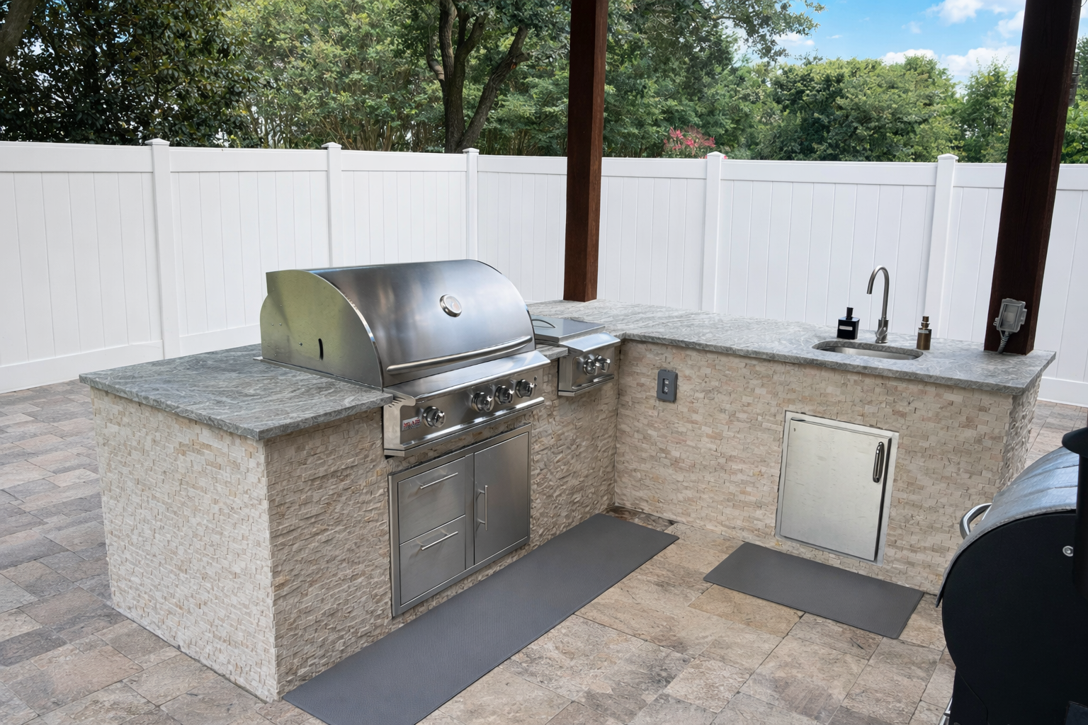 Outdoor kitchen with stainless steel grill, stone countertop, sink with soap dispenser, paper towel holder, and a small cabinet, surrounded by a white fence and trees in the background.