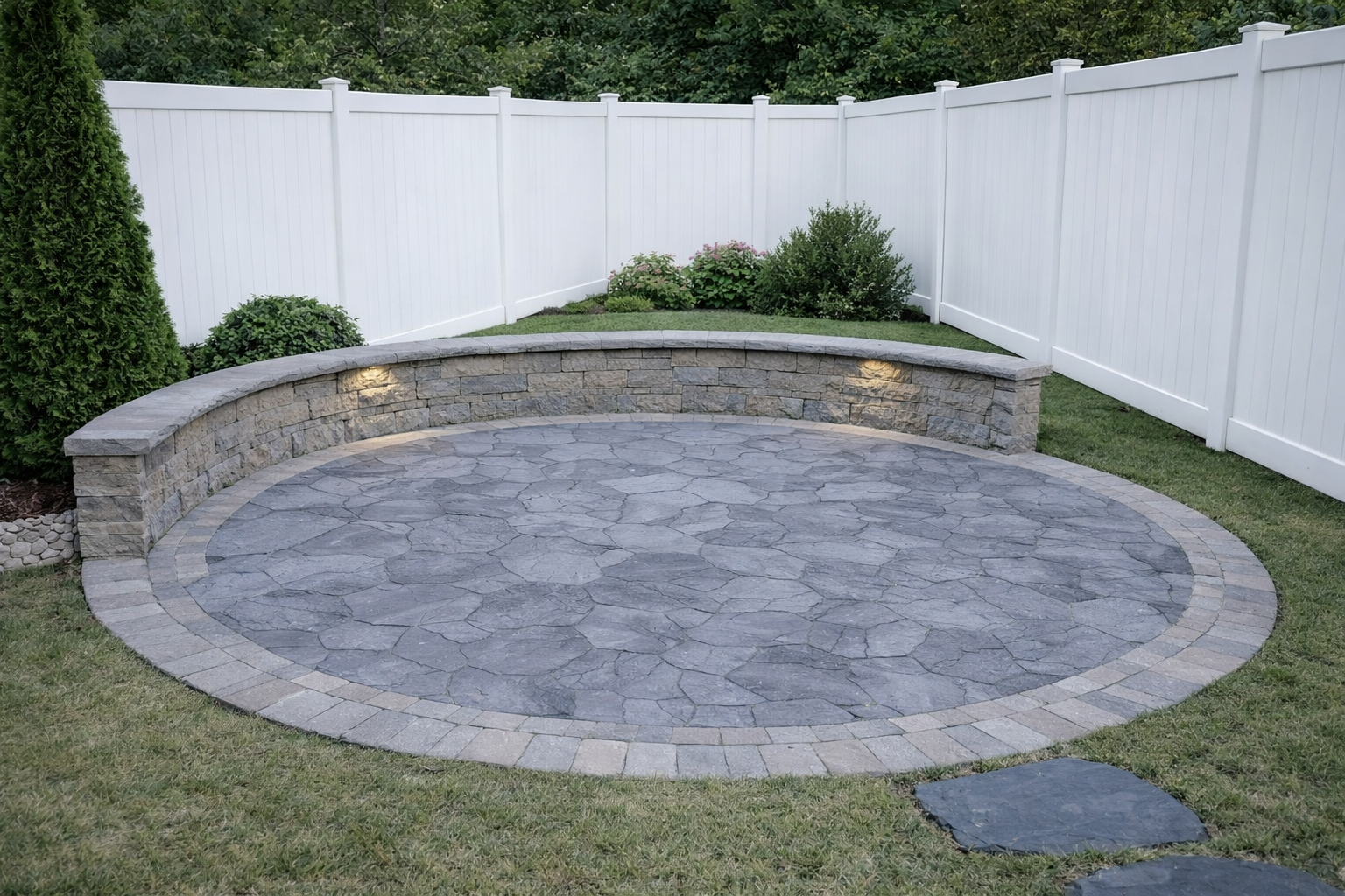A circular stone patio with a curved stone wall, surrounded by grass, bushes, and a white privacy fence, with outdoor lighting in the wall.
