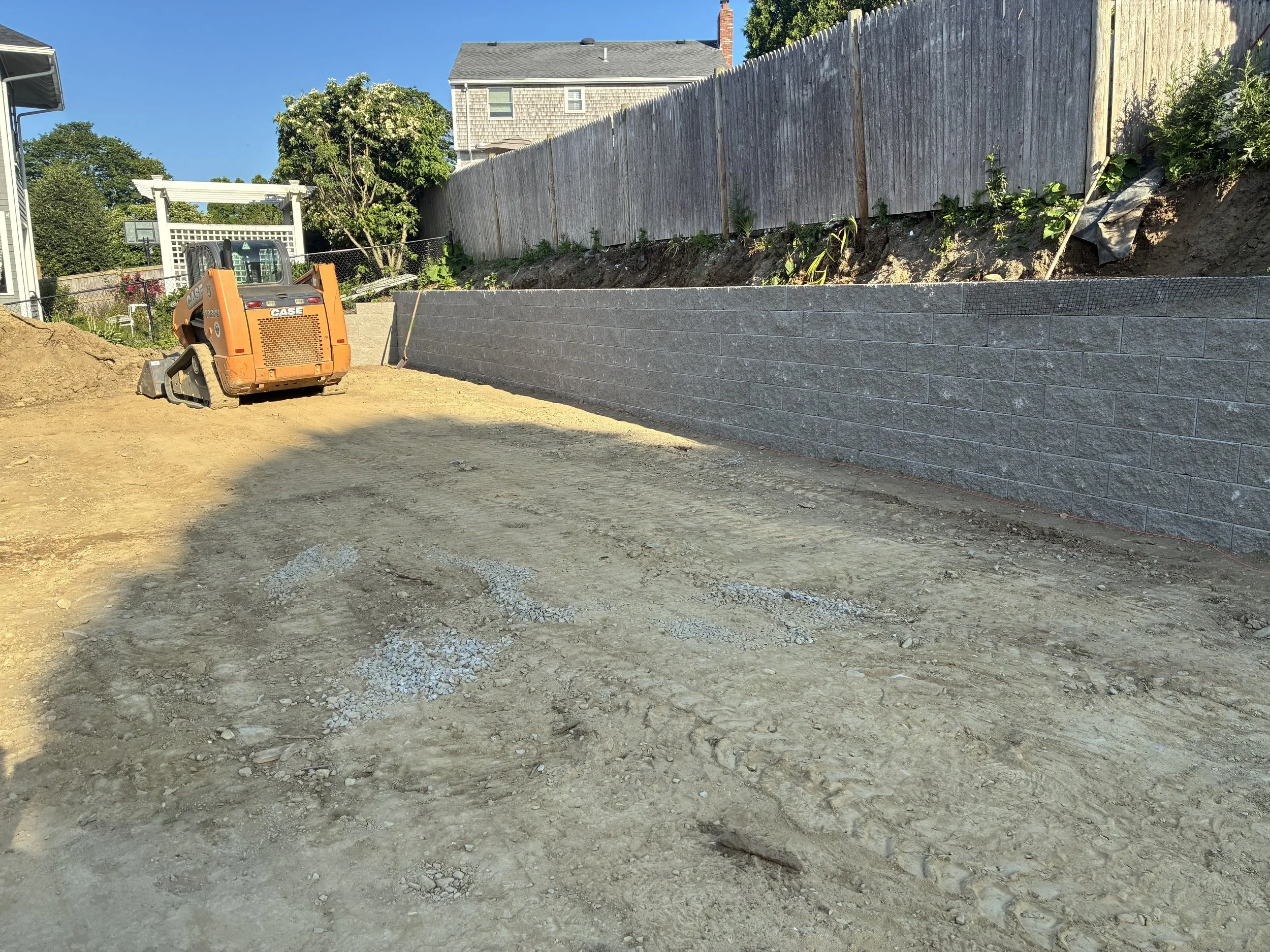 Construction site with a dirt ground, a small orange excavator, and a new gray brick retaining wall along a slope behind a wooden fence.