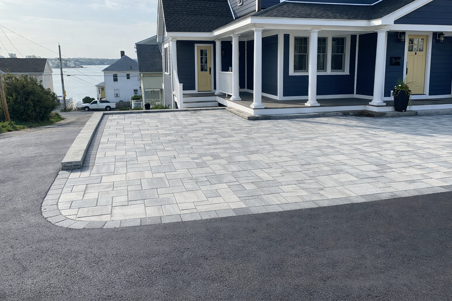 A newly paved driveway with grey interlocking bricks in front of a blue house with white trim and yellow doors, overlooking a body of water with neighboring houses in the background.