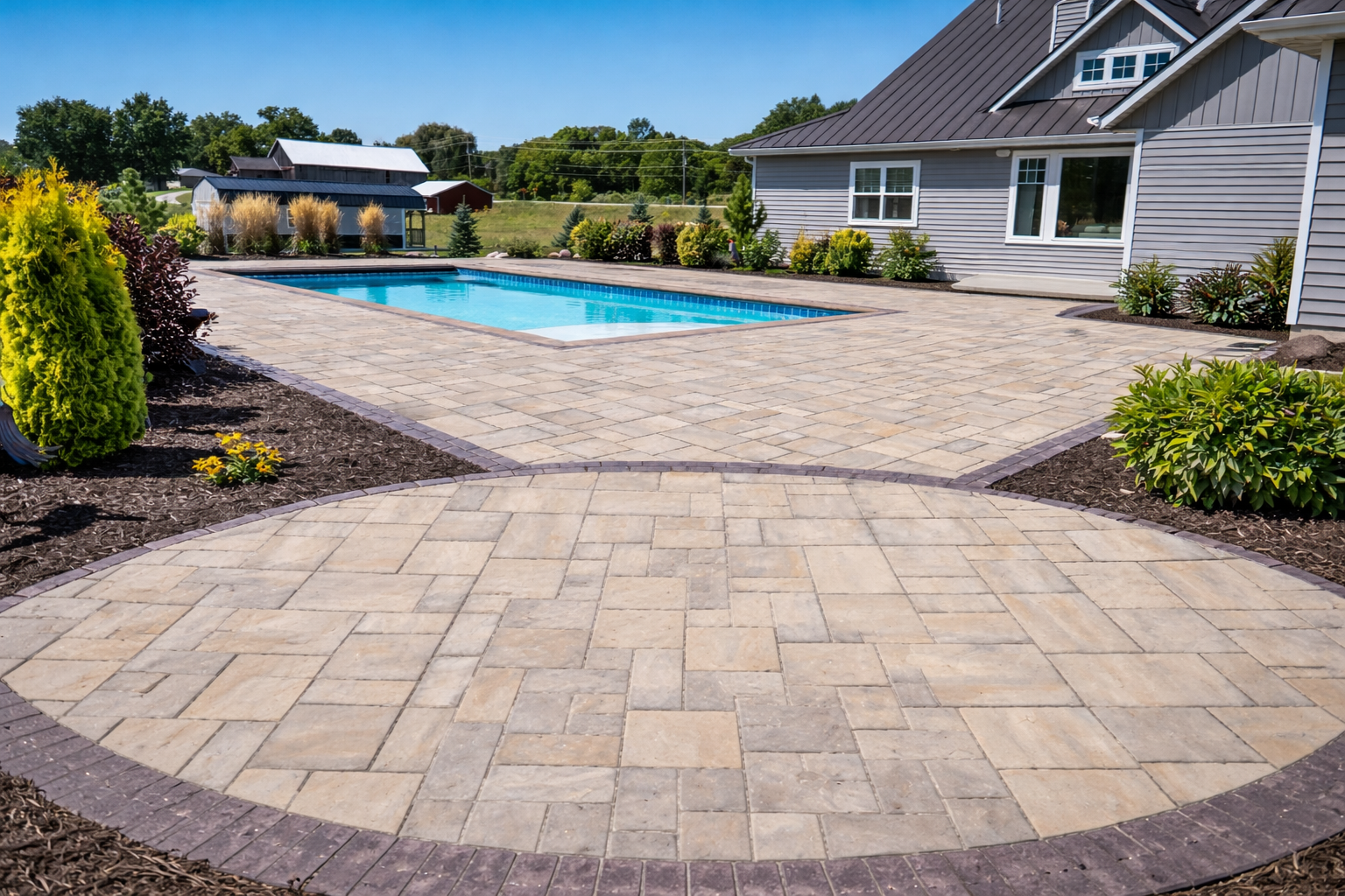 A backyard with a rectangular swimming pool, a paved patio, and landscaped garden beds with bushes and plants, next to a house with gray siding and large windows.