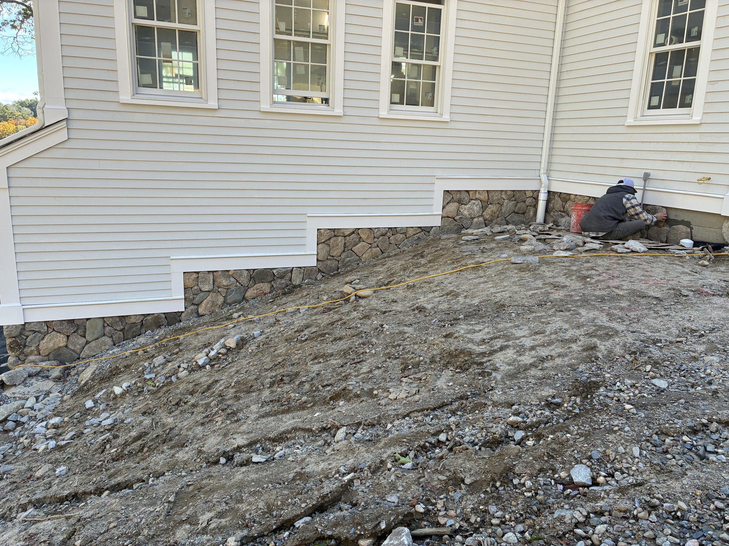 A worker is kneeling and applying mortar or cement to the foundation of a house, which has white siding and stone work at the base.