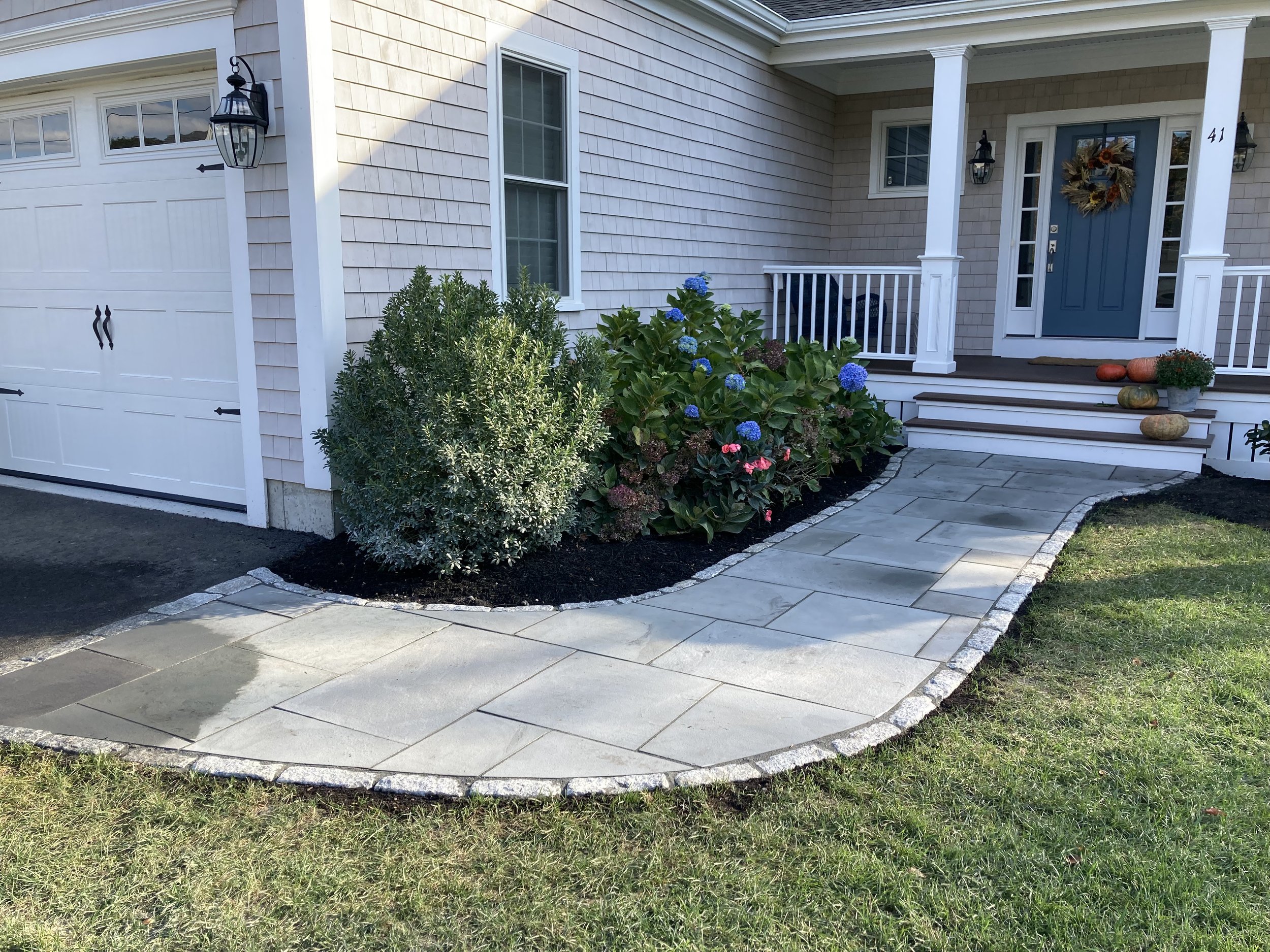 A front porch with a concrete pathway leading to steps in front of a house with a blue door decorated with a sunflower wreath. The porch has white railings, and there are pumpkins and potted plants on the steps. There are shrubs and flowering plants 