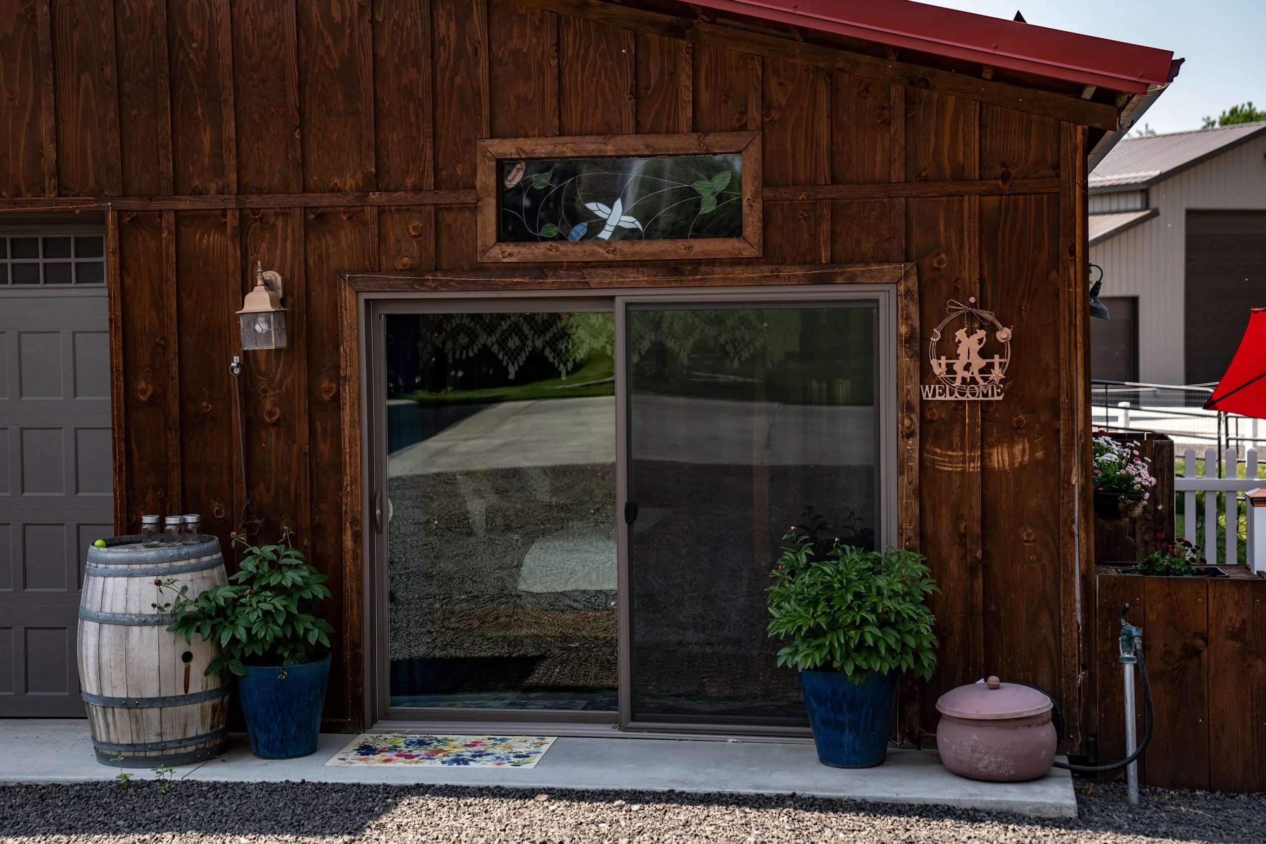 Front of a wooden barn with sliding glass door, potted plants, a barrel, a pink ceramic container, a welcome sign, and outdoor lighting fixtures.