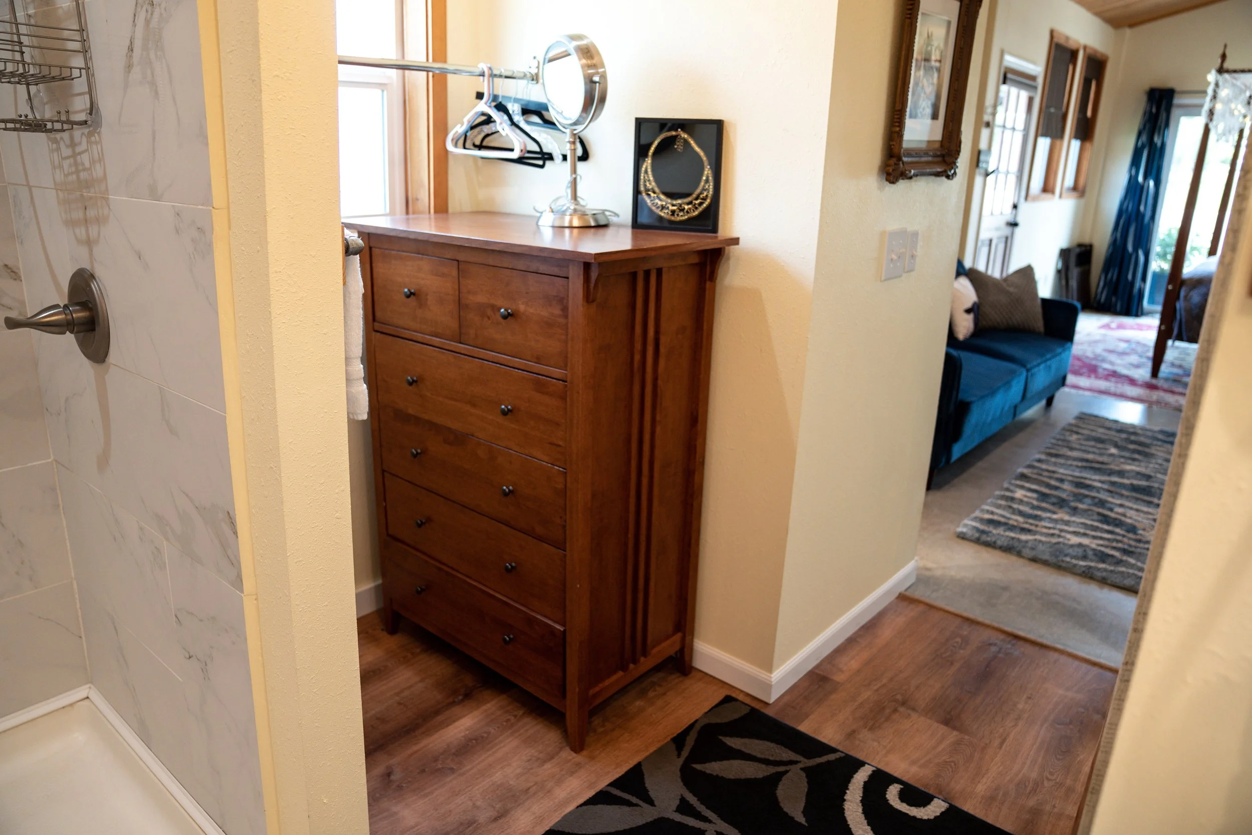 Interior view of a home showing a wooden dresser near a hallway, with a jewelry stand and a small picture frame on top. The hallway leads to a living room with a blue couch and patterned rugs, and a door to the outside visible in the background.