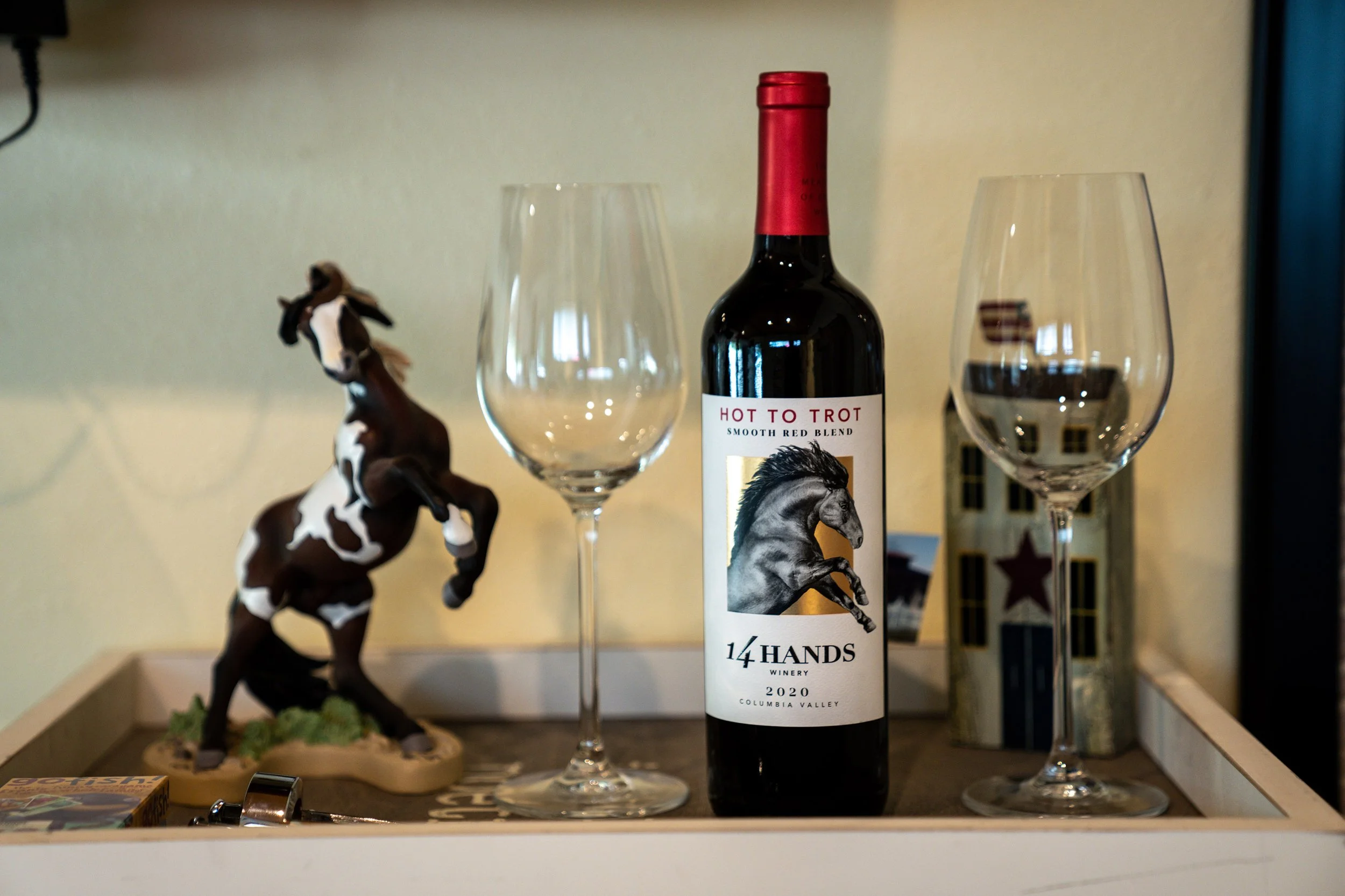 A bottle of red wine labeled 'Hot to Trot, Smooth Red Blend, 14 Hands Winery, 2020, Columbia Valley' stands between two empty wine glasses. To the left, there is a decorative horse figurine, and to the right, a lantern with star-shaped cutouts. A sma