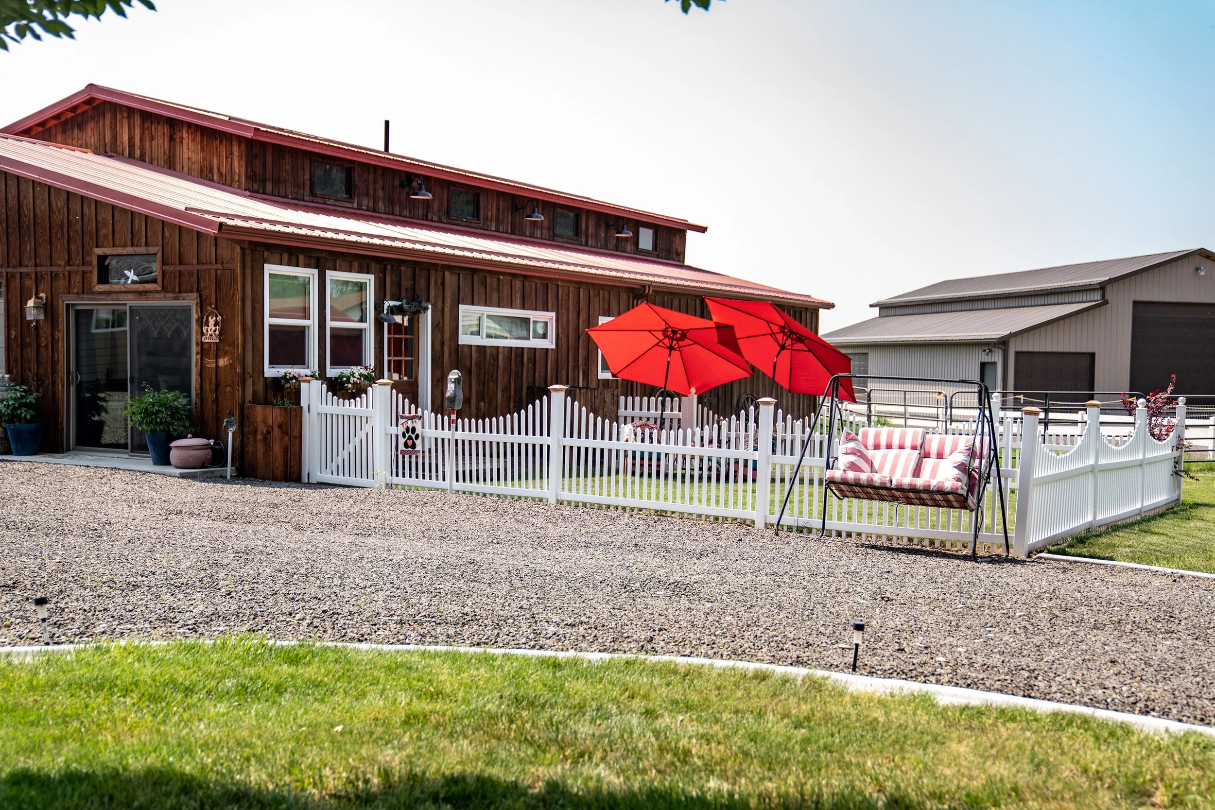 A rustic wooden barn with a red metal roof, a white picket fence, two red umbrellas, and a striped swing outside.