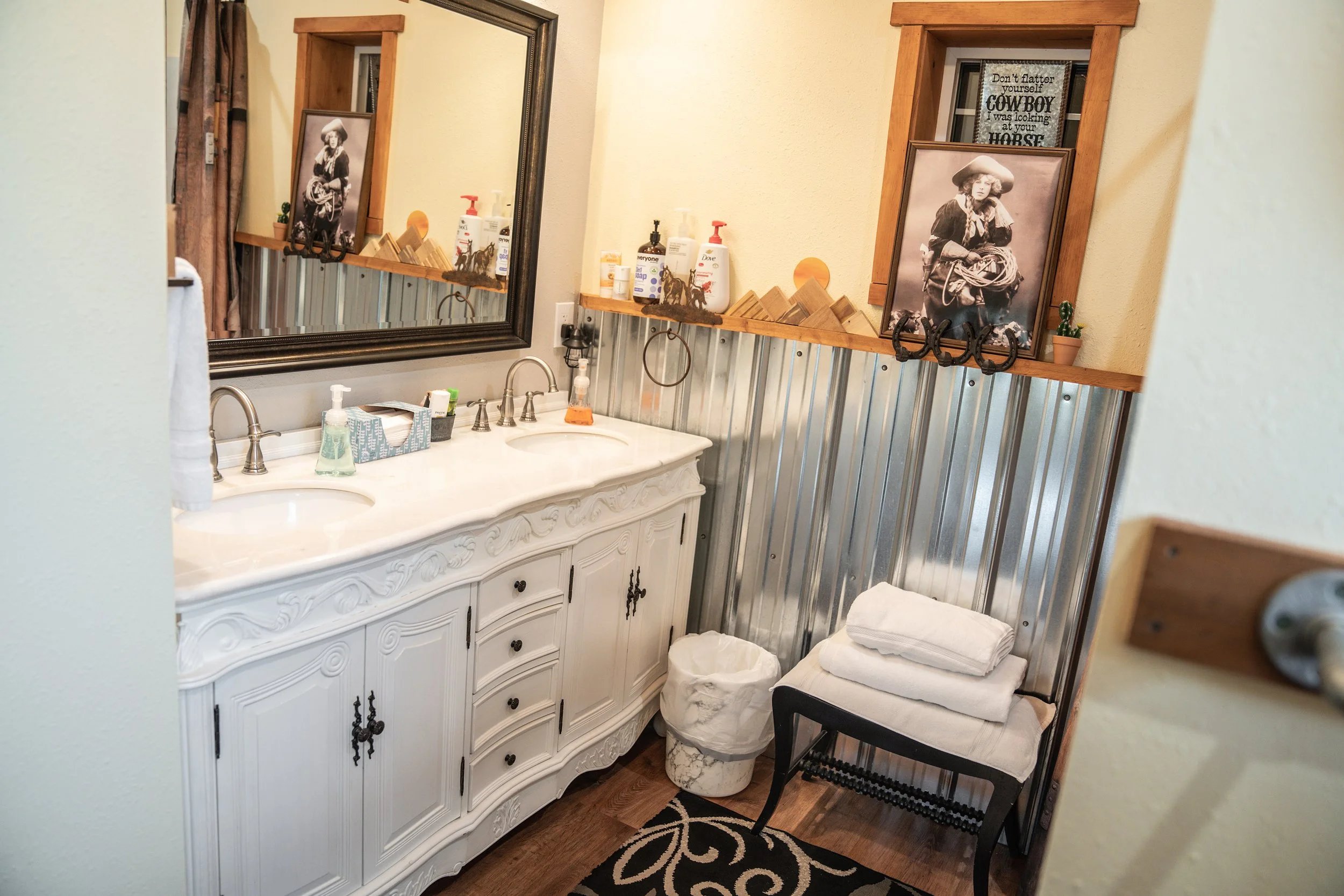 Bathroom with a double sink vanity, large mirror, framed black and white portraits, decorative wood and metal accents, towels, toiletries, trash bin, and a small black table with folded towels.