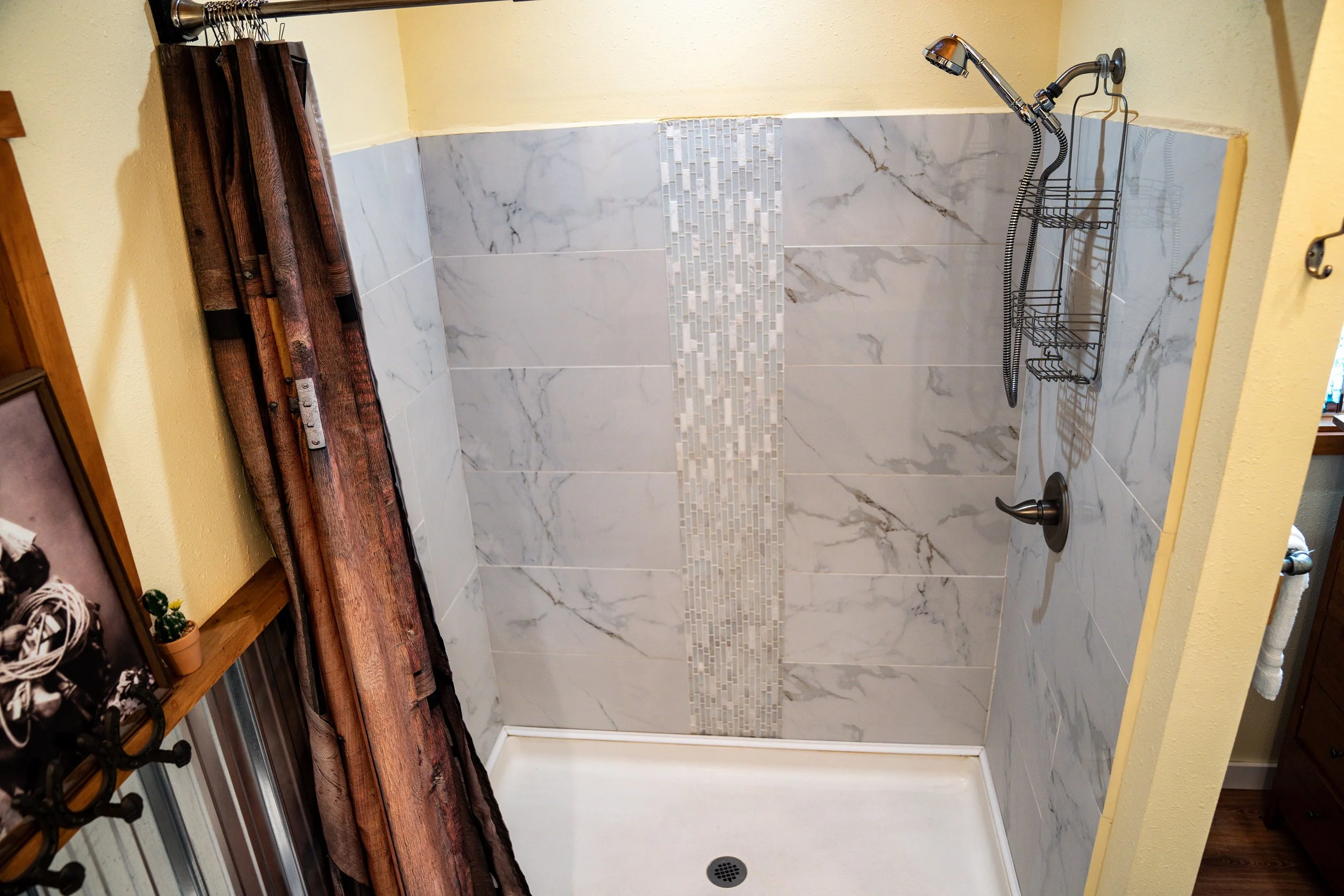 Empty shower with marble tile walls and a tiled vertical strip in the center, a showerhead, a wire soap holder, and a small drain in the white shower pan.