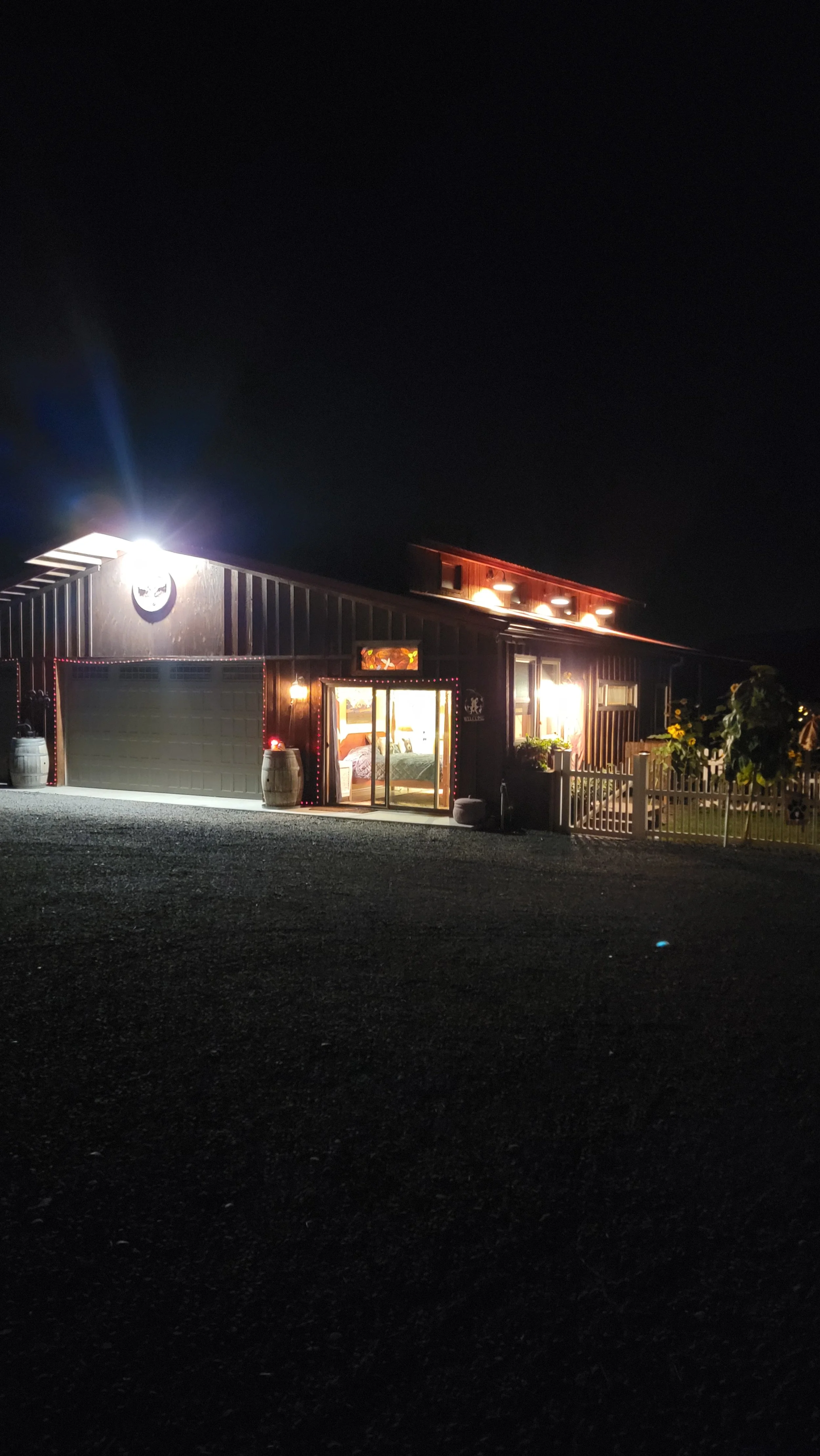 Nighttime view of a rustic house with warm interior lighting and decorative string lights outside, with a gravel driveway in the foreground.