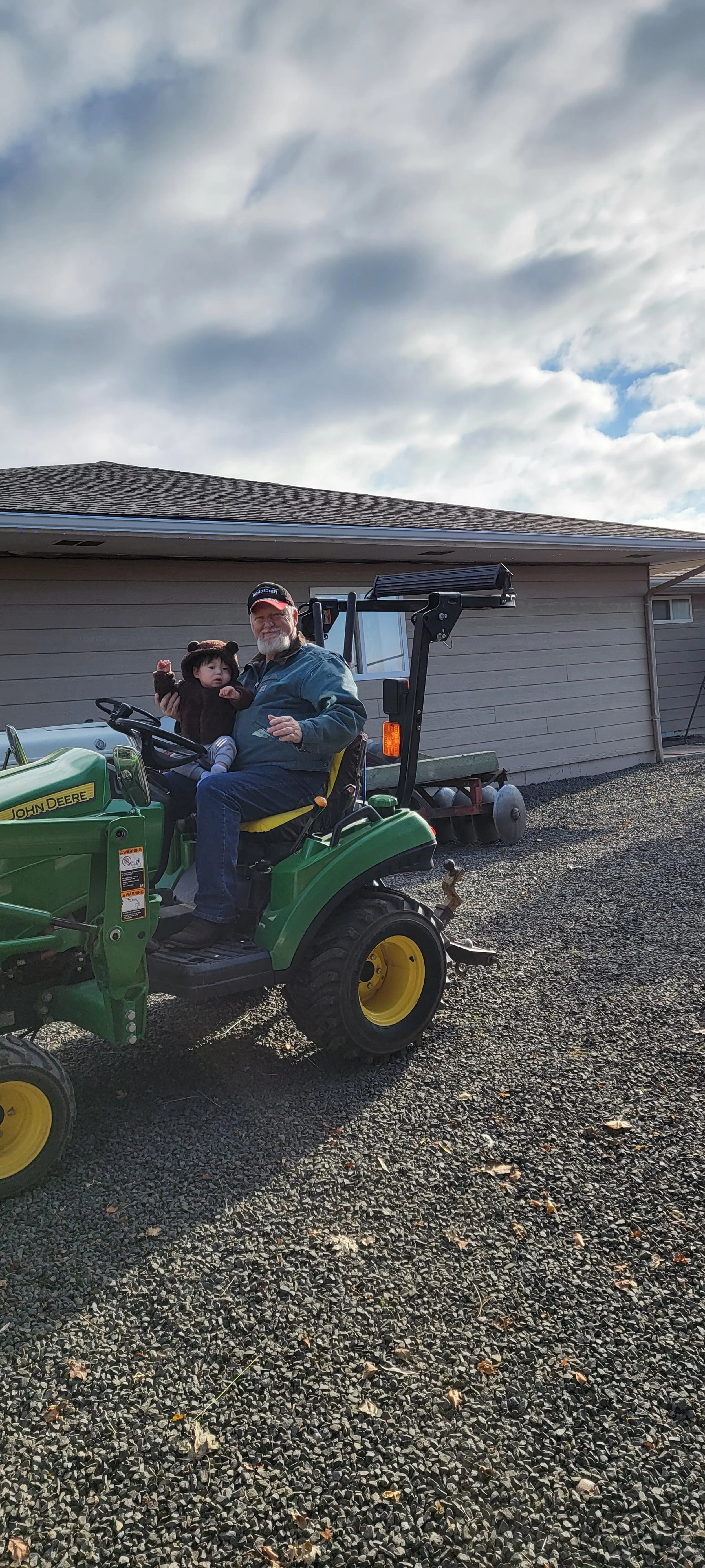 A man with a child sitting on a green John Deere tractor parked on a gravel driveway in front of a house with beige siding.
