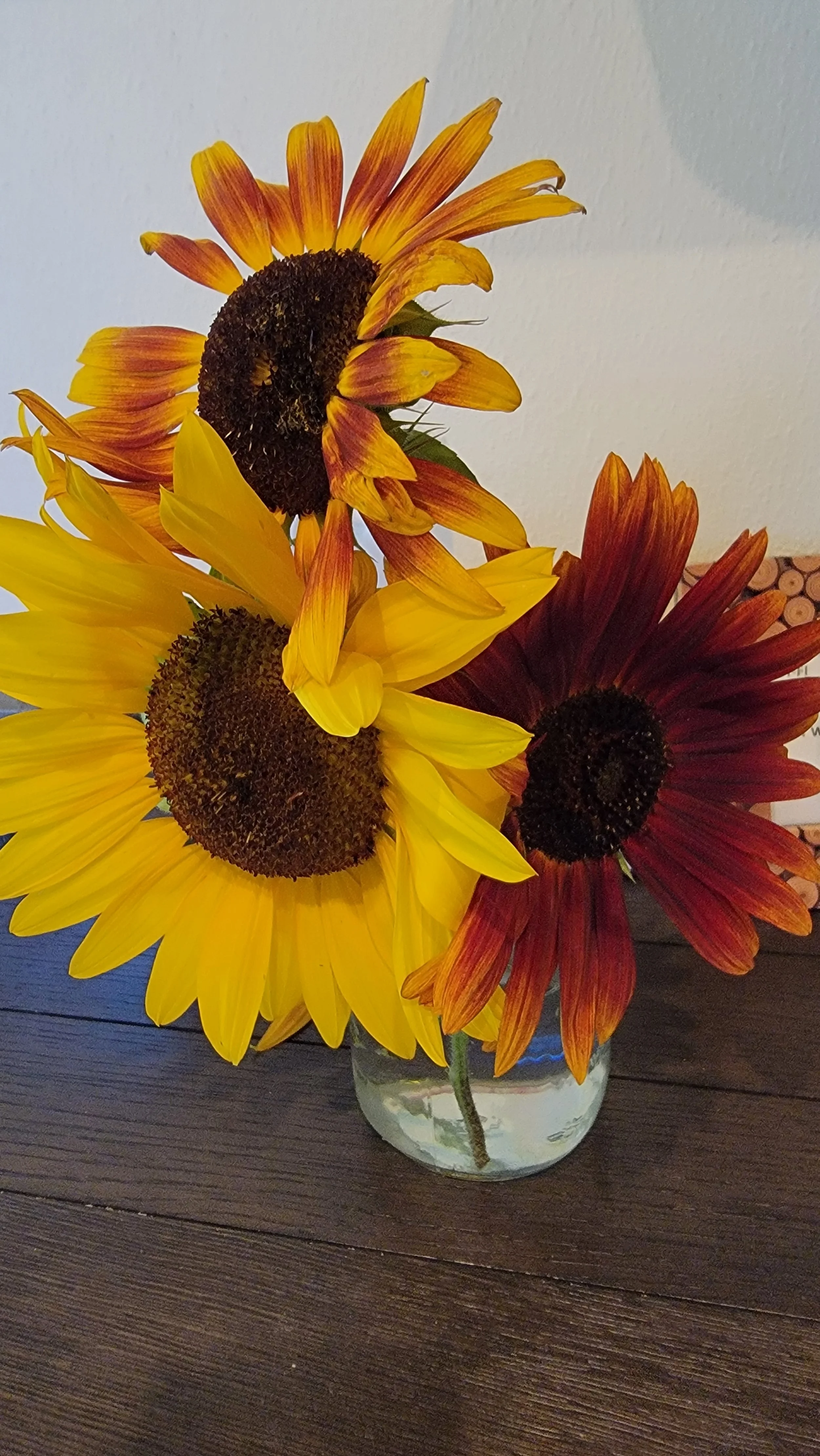 A bouquet of three sunflowers in a glass vase on a dark wooden table, against a plain wall background.
