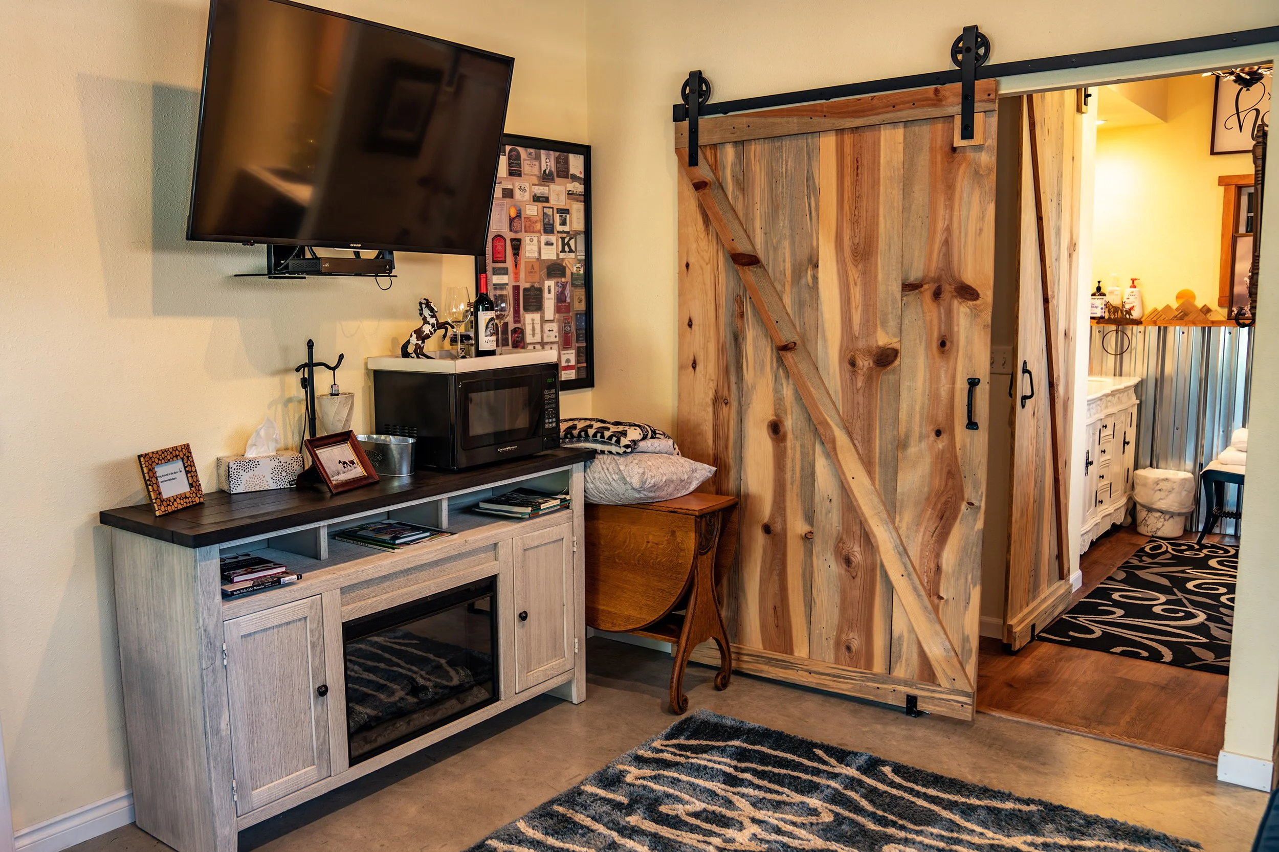 A rustic wooden sliding barn door partially open, revealing a bathroom with a sink, countertop, and various toiletries. Inside the room, there is a white wooden cabinet with a mirror above, a black and white rug, and various decorative items.