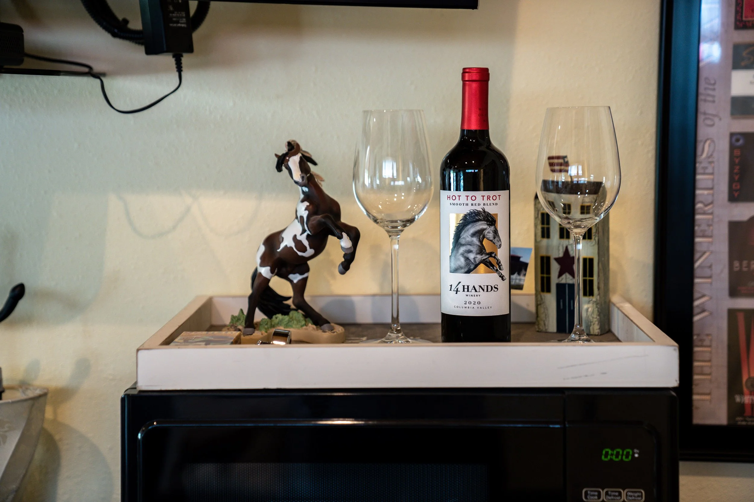 A decorative tray on top of a microwave oven holding a small horse figurine, a bottle of red wine, two empty wine glasses, and a photo frame.