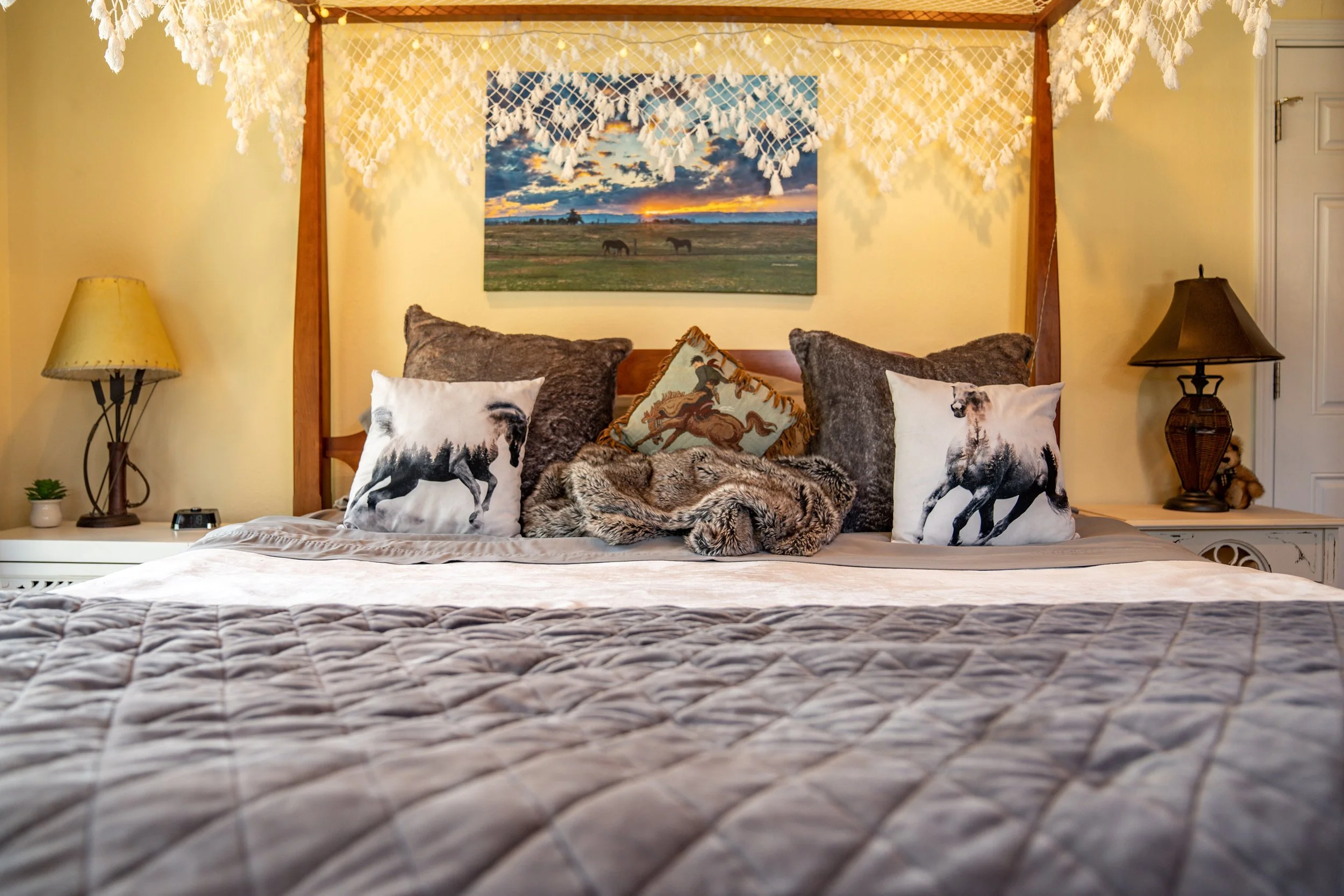 A cozy bedroom with a four-poster bed adorned with forest animal-themed pillows, animal print throws, and a furry blanket. The bed has a landscape painting of horses and sunset above it. Two bedside tables with lamps, one with a small plant and a ted
