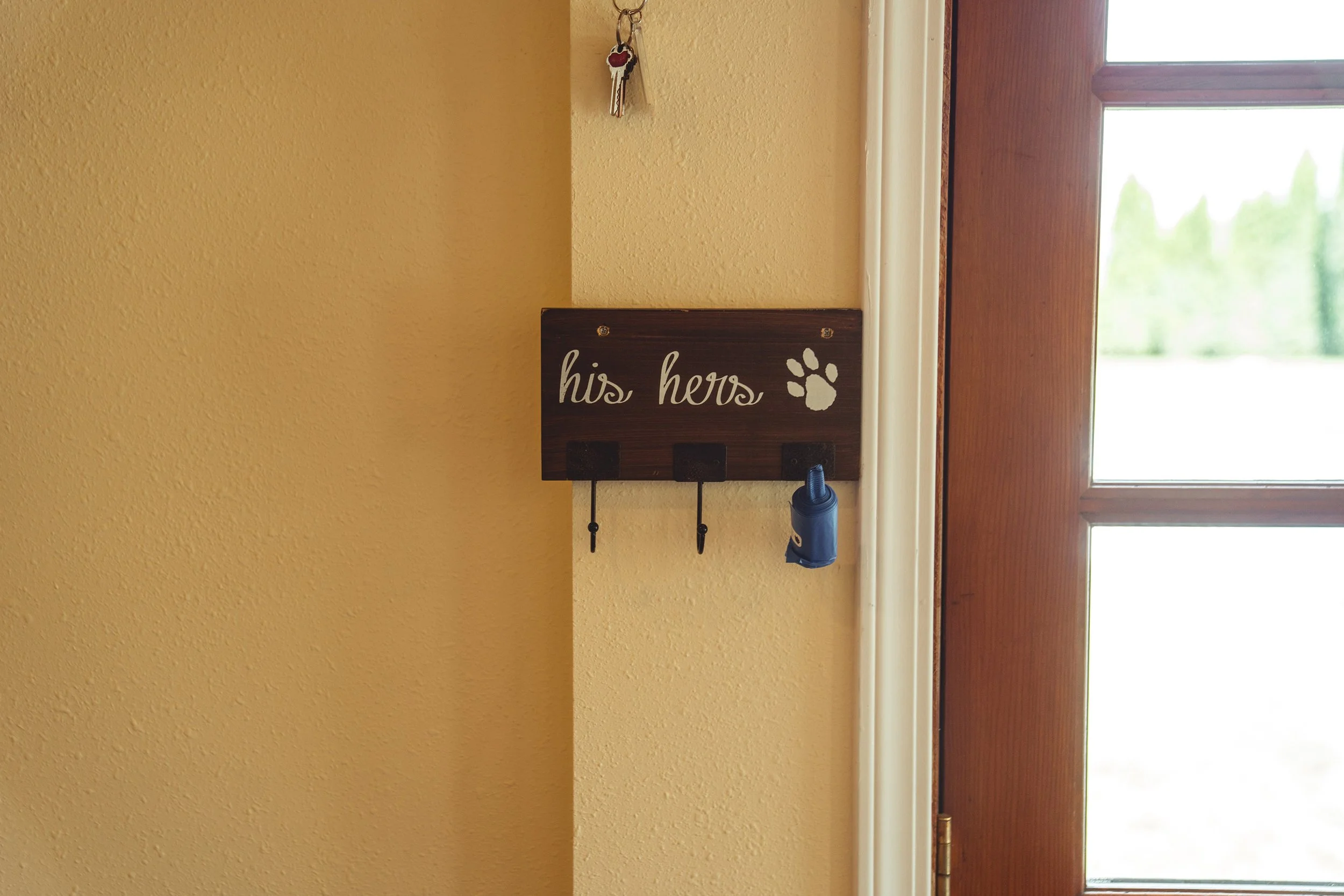 A wooden wall hook rack labeled 'his hers' with paw print, mounted next to a door with a window, holding a blue dog waste bag and keys.