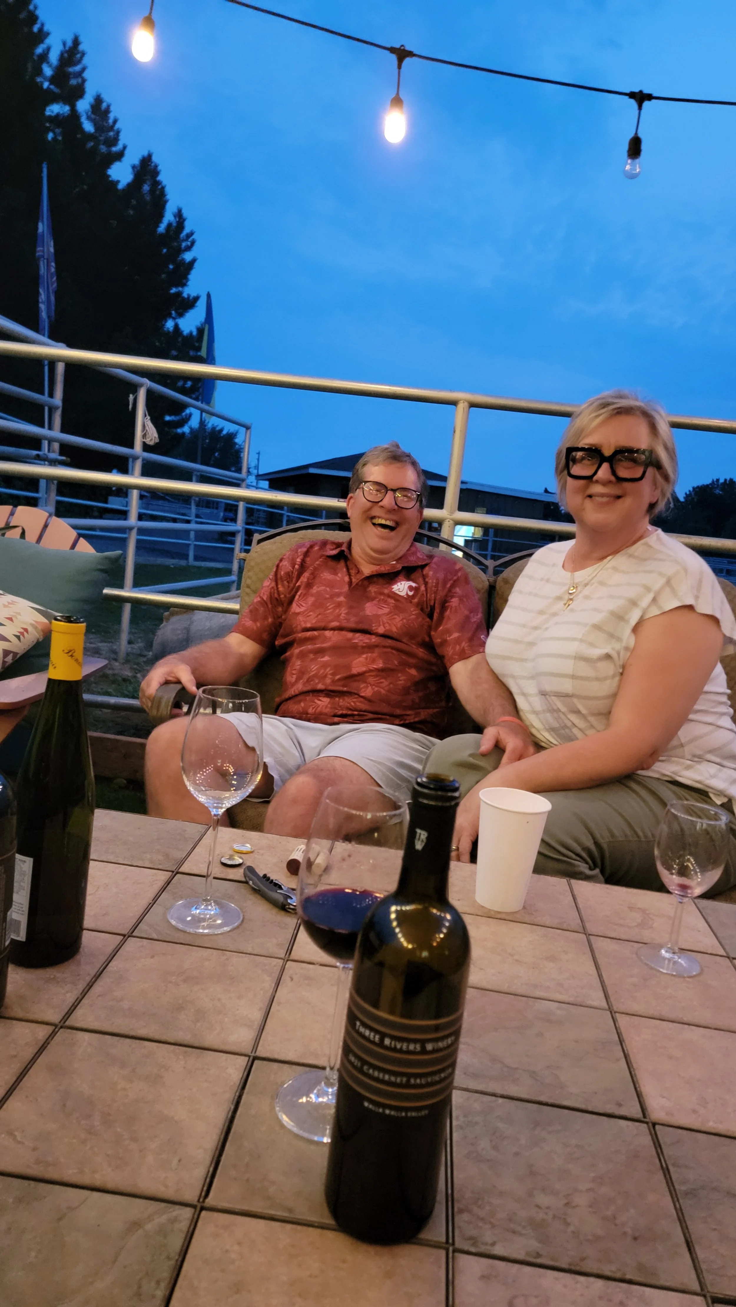 Two smiling people, one man and one woman, sitting outdoors at a table with drinks, under string lights at dusk.